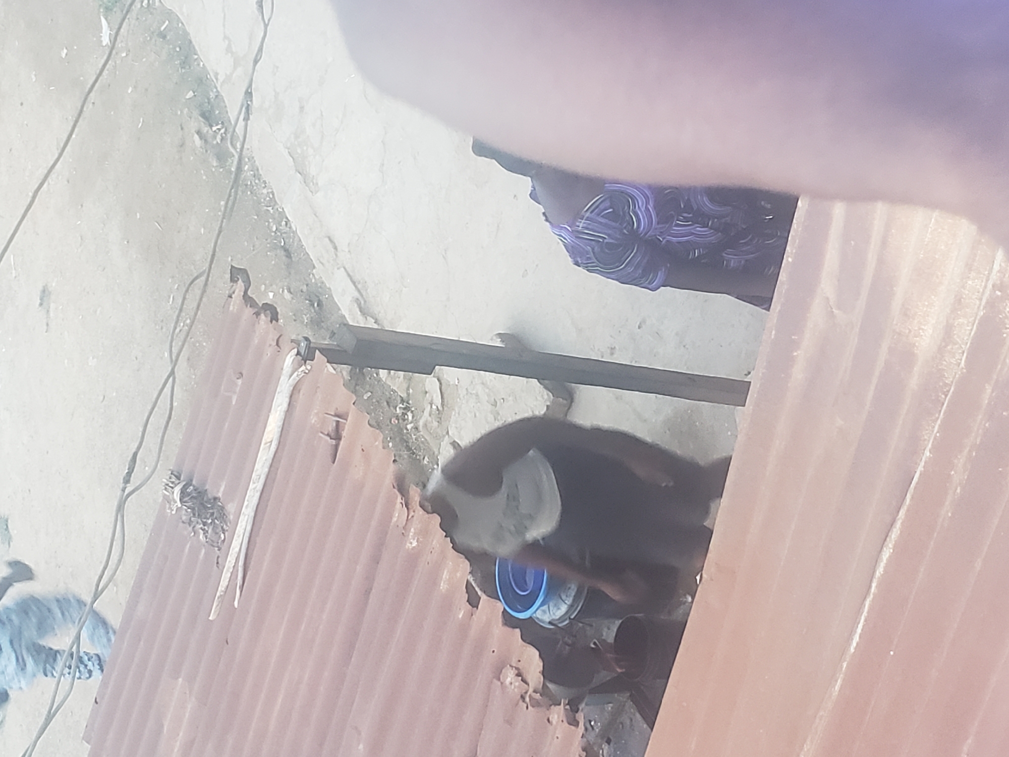 The image shows a scene in Alalubosa, Nigeria. A person wearing a dark patterned shirt is partially visible near a section of corrugated metal. A black and white goat is also visible, standing near a blue bucket. The background appears to be a concrete or stone surface.