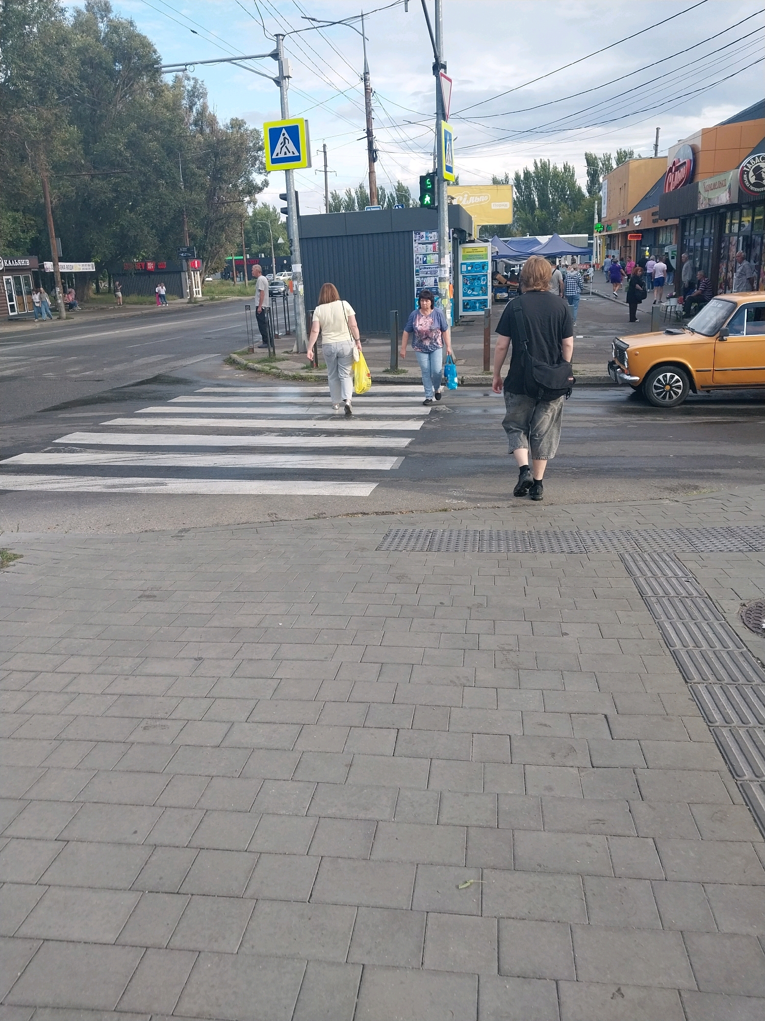 The image depicts a street scene in Dnipro, Ukraine, likely on a pedestrian crossing near commercial establishments. In the foreground, a person with light brown hair wearing a black t-shirt and grey shorts walks away from the camera, crossing a paved sidewalk. The sidewalk is made of grey rectangular pavers, with a tactile paving strip to the right. The pedestrian crossing itself is clearly marked with white stripes across the asphalt road. Two women are walking on the pedestrian crossing, one in the foreground to the left wearing a white top and light grey pants, carrying a yellow bag, and another further back wearing a blue patterned top and jeans, carrying a blue bag. Behind them, a man in a white shirt and dark pants stands on the sidewalk. On the right side of the road, an orange vintage car is parked. In the background, there are several shops with awnings and signs, suggesting a commercial area. A pedestrian crossing sign with a blue background and white figures is visible on a pole. Trees line the left side of the road, and overhead wires crisscross the sky. The sky is overcast with a mix of blue and grey clouds.