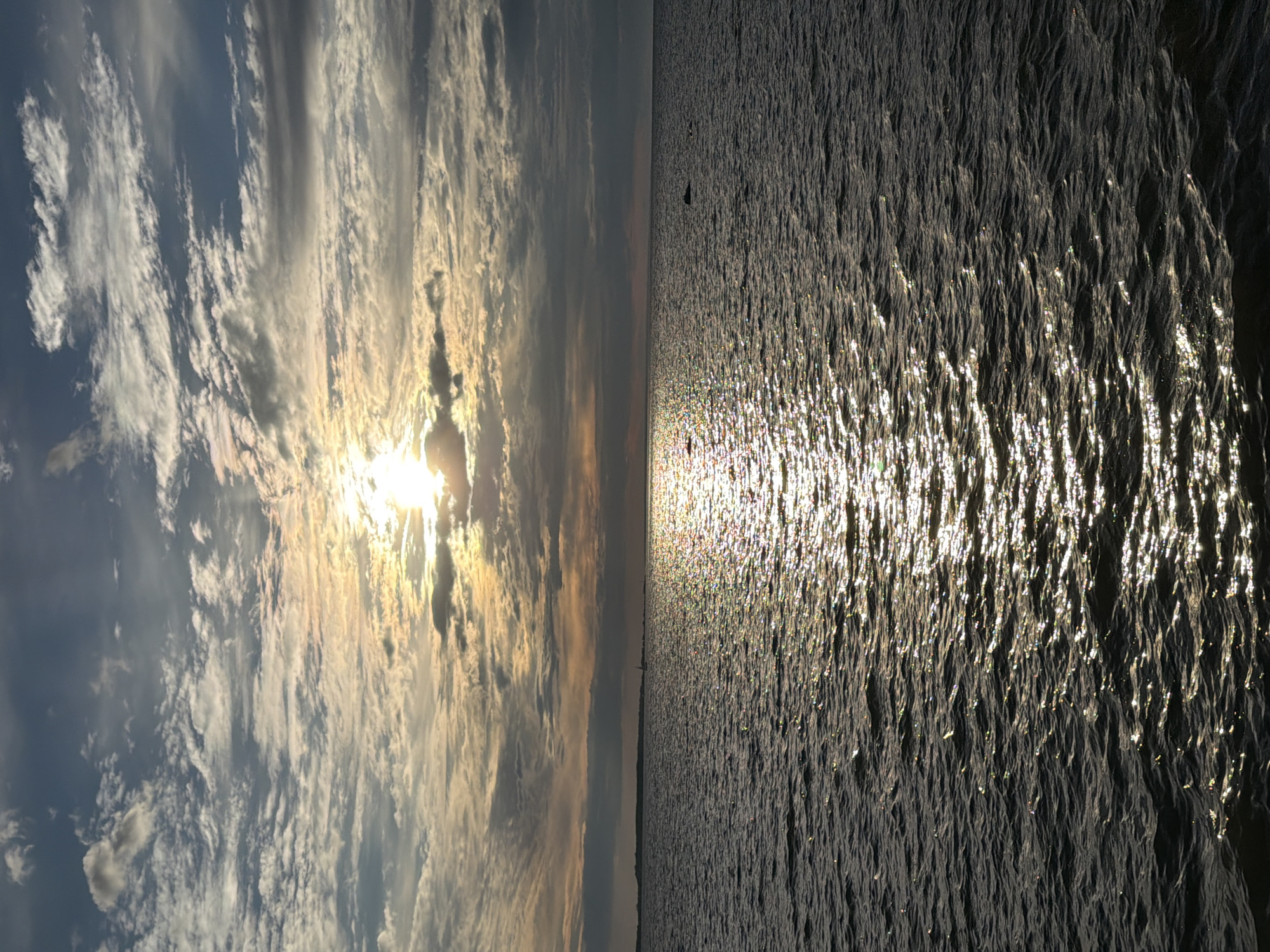 The image depicts a body of water, likely the sea, under a cloudy sky during what appears to be sunset or sunrise. The sun is partially obscured by clouds, casting a bright glow that reflects off the water's surface, creating a shimmering path of light. The water is calm with gentle ripples. The horizon is visible in the distance, with a faint silhouette of land and possibly a sailboat. The sky is a mix of blue and gray clouds, with streaks of orange and yellow indicating the sun's presence. The overall scene is serene and atmospheric. This image could have been taken from the coast of Tallinn, Estonia, looking out towards the Baltic Sea.