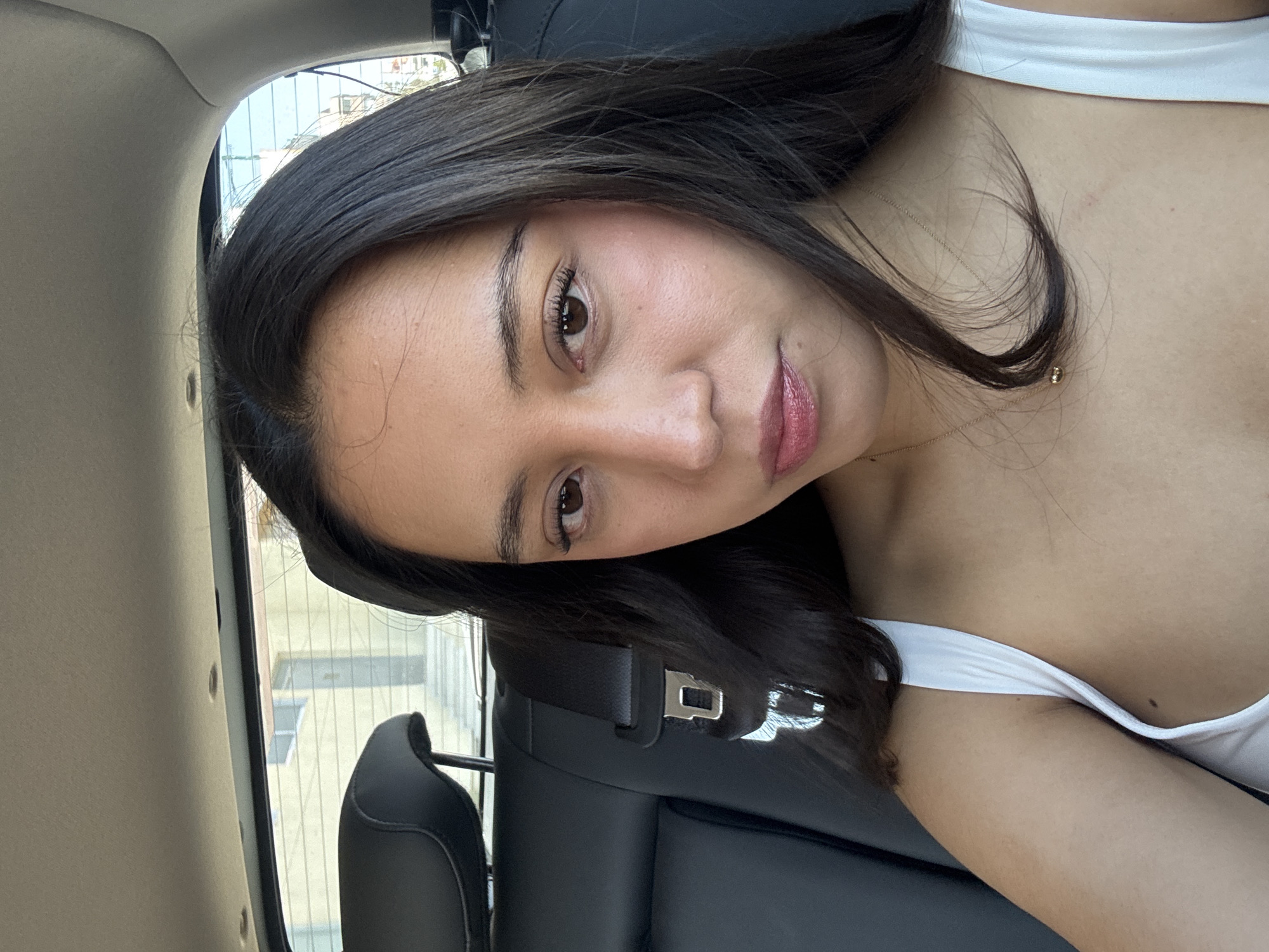 A young woman with long, dark hair is sitting in a car, wearing a white top. She is wearing a seatbelt. The background shows buildings, possibly indicating an urban area. The image does not provide enough information to pinpoint the location to the city of Puebla, Mexico.