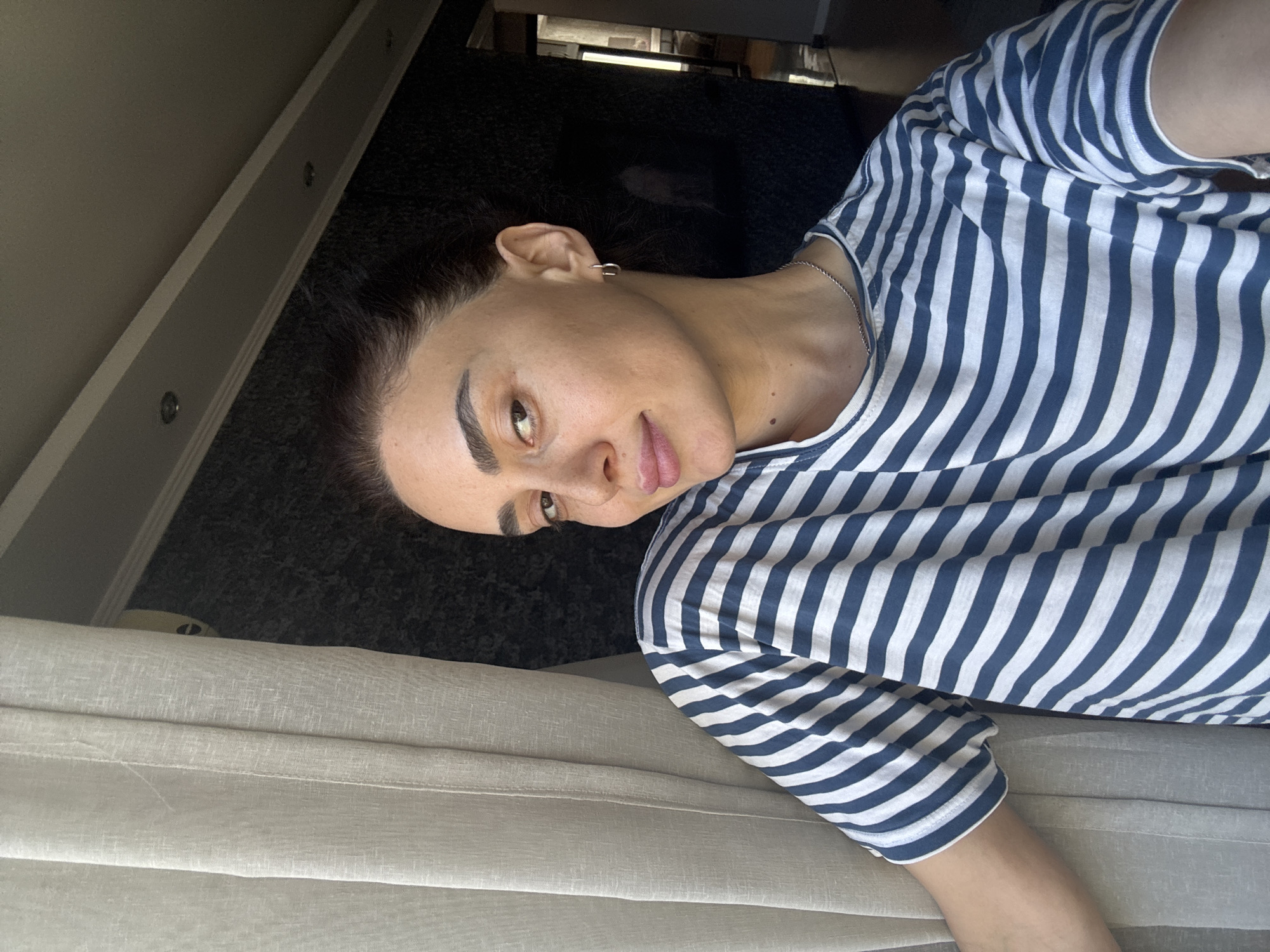 A medium shot captures a woman with dark hair pulled back, her face turned towards the camera with a slight smile. She is wearing a blue and white striped t-shirt and a silver necklace. A light-colored curtain hangs to her left, partially obscuring her arm. The background features a dark, textured wall and a ceiling with recessed lighting. The lighting suggests an indoor setting, possibly a home in Tbilisi, Georgia, given the context.