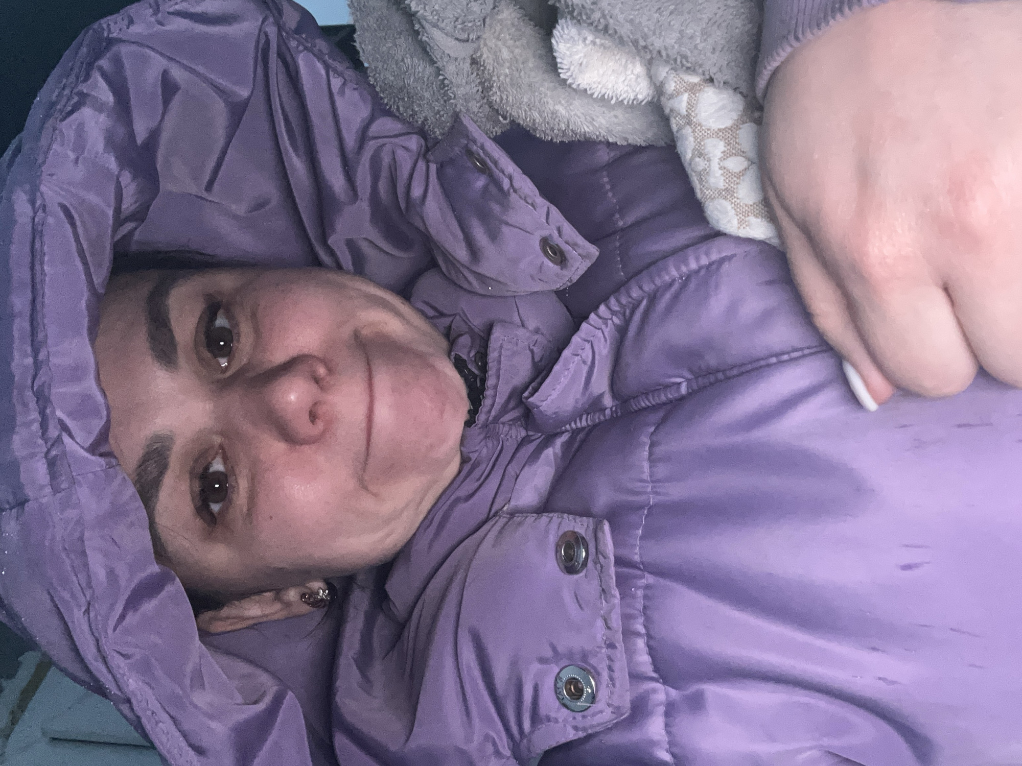 A woman is lying down, wearing a purple hooded jacket. A hand and a towel are visible next to her. The location is Chaikovsky, Russia.