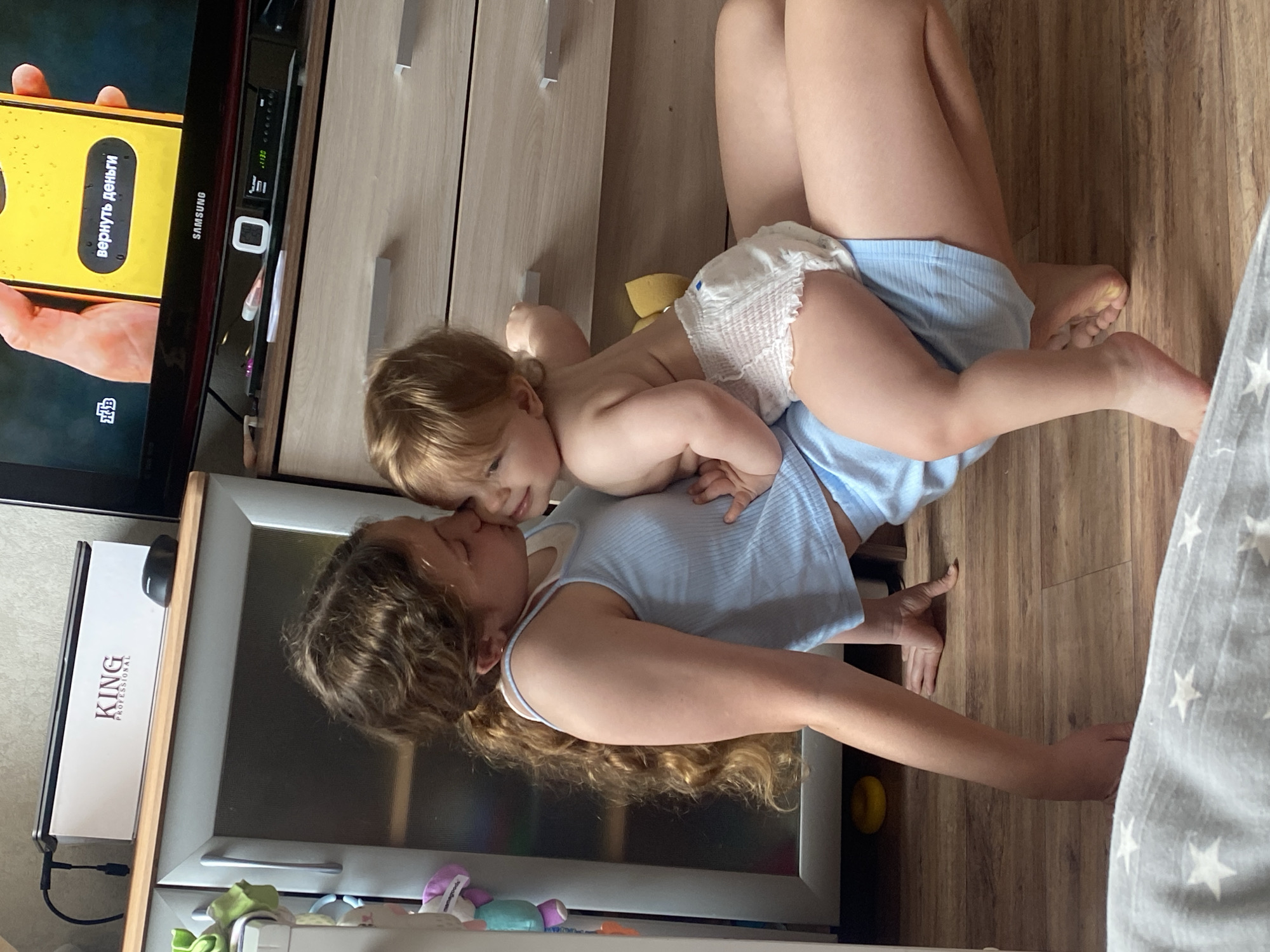 An overhead shot shows an adult woman lying on her back on a wooden floor, with a young child (likely a toddler) lying on top of her, face-to-face. The woman has long, curly brown hair and is wearing a light blue ribbed top. Her eyes are closed, and she appears relaxed. The child, with fair hair and wearing a white diaper and light blue shorts, is looking towards the woman, their faces very close, suggesting an affectionate interaction like cuddling or playing.
The setting is an indoor space, possibly a living room or bedroom. To the left, a television screen is visible, showing a yellow mobile phone display. On the phone screen, Russian text reads "ВЕРНУТЬ ДЕНЬГИ" (VERNUT' DEN'GI), meaning "RETURN MONEY," and "НТВ" (NTV) is in the corner. The TV itself has the brand "SAMSUNG" visible. Above the TV, a light wood cupboard is seen.
To the bottom left, next to the woman's head, there's a reflective surface, possibly a mirror or the door of a refrigerator, with a white box labeled "KING PROFESSIONAL" on top. Below this surface, some colorful soft toys or accessories are visible. The wooden floor continues across the image, and a gray fabric with white stars is partially visible on the right, likely part of a blanket or furniture. A small yellow object is on the floor near the woman's right arm. The overall atmosphere conveys warmth and familial bonding.