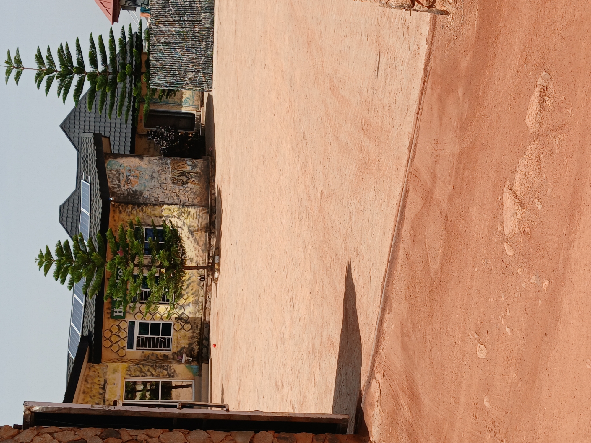 The image shows a side view of a building in Kwang, Nigeria. The building has a distinctive architectural style with a dark tiled roof and several windows. Solar panels are visible on the roof, suggesting an eco-friendly setup. The walls of the building are adorned with colorful murals and graffiti, giving it a unique and artistic appearance. A tree grows in front of the building, adding a touch of nature to the scene. The ground in front of the building is covered with a sandy, reddish-orange surface, typical of some Nigerian landscapes. A distinct shadow of the tree is cast on the ground, indicating it's likely daytime, with the sun at an angle. The overall impression is of a vibrant and culturally rich location. There is visible text on the building that appears to be a logo, possibly "Heineken". The scene evokes a sense of local artistry and everyday life.