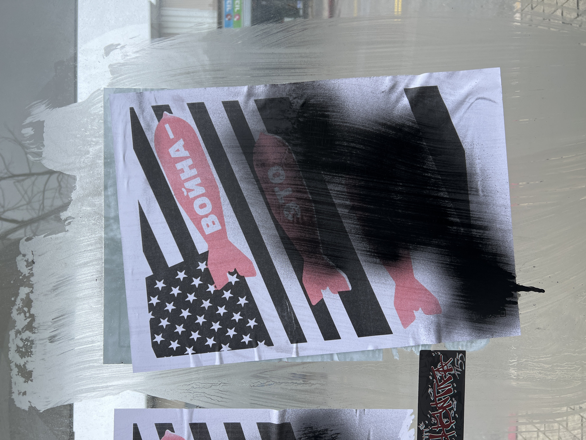 The image displays a poster adhered to a smudged glass surface, likely a bus stop or storefront window, in an urban setting. The poster features a stylized rendition of the American flag, with black and white stripes and a black canton containing white stars. Replacing the red stripes are several pink, bomb-shaped silhouettes. The uppermost pink bomb contains the Russian word "ВОЙНА" (VOYNA), meaning "WAR," in white capital letters. The lower half of the poster is heavily obscured by black spray paint, which shows drips at the bottom edge. To the left, a portion of an identical or similar poster is visible. Below and to the left of the main poster, a smaller black rectangular sticker or tag with red and white graffiti-style lettering and the number "1155" is affixed. Through the glass, blurry outdoor elements are discernible, including what appears to be trees and a faint, colorful sign in the background. The precise location within Tel-Aviv cannot be determined from the visible details.