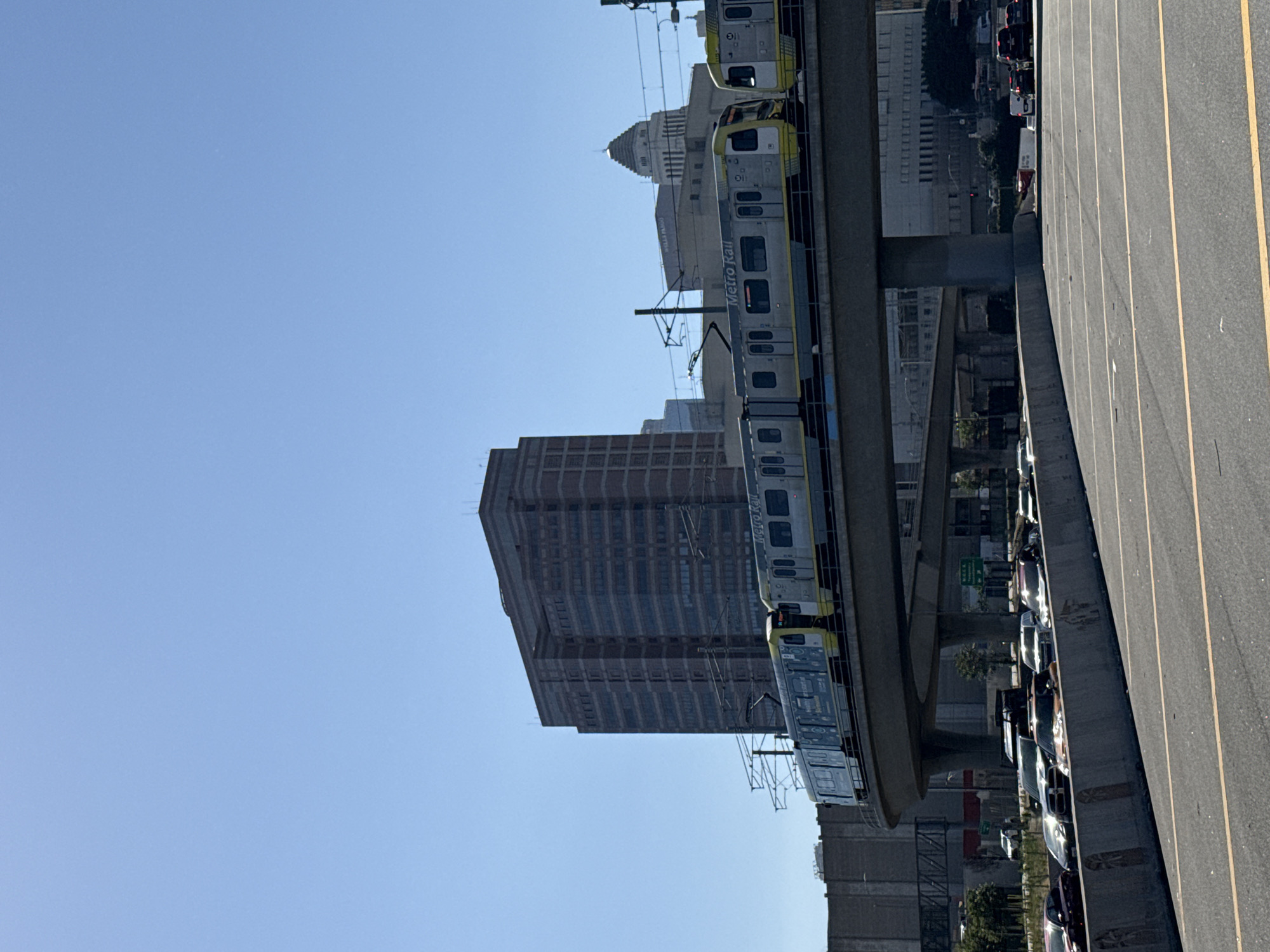 The image depicts a scene in Los Angeles, California, featuring a Metro Rail train on an elevated track. The train cars are light gray with yellow accents and display the "Metro Rail" logo. In the background, there are several buildings, including a tall, brick-clad building and a shorter, lighter-colored building with a dome. To the right of the train track, a busy road is visible with multiple lanes of traffic. Cars of various colors and models are lined up. The sky is a clear, light blue.