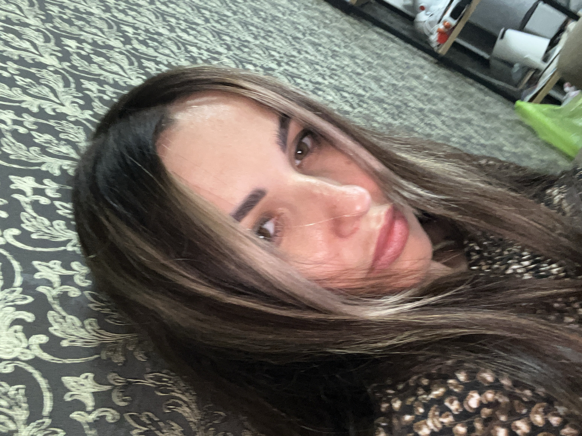 A woman is looking at the camera in a close-up selfie. She has long, wavy, dark brown hair with blonde highlights. Her eyes are brown, and she has full lips. She is wearing a dark patterned top. The background is a wall with a textured, floral pattern and to the right, a shelf with items on it, including shoes and a white container. The image is taken in an indoor environment.