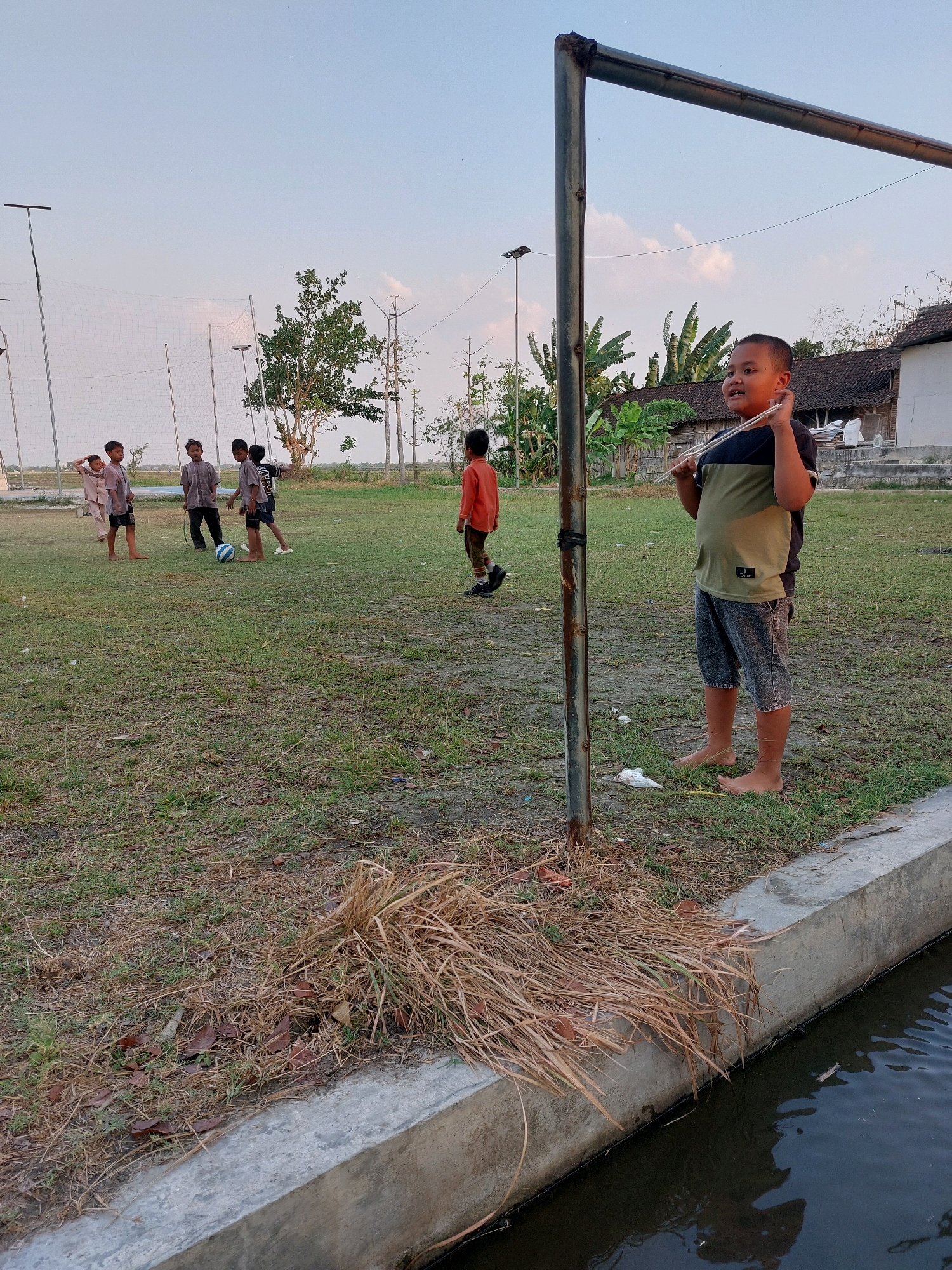 In Sarangan, Indonesia, a group of young boys are playing soccer on a grassy field. A boy in the foreground, wearing a black and green t-shirt and denim shorts, stands near a goalpost holding a makeshift toy. Other boys are scattered across the field, with one boy near the center of the frame kicking a soccer ball. The field appears to be part of a recreational area, with a concrete structure and water body bordering one side of the field. The sky is clear and a few trees and buildings are visible in the background.