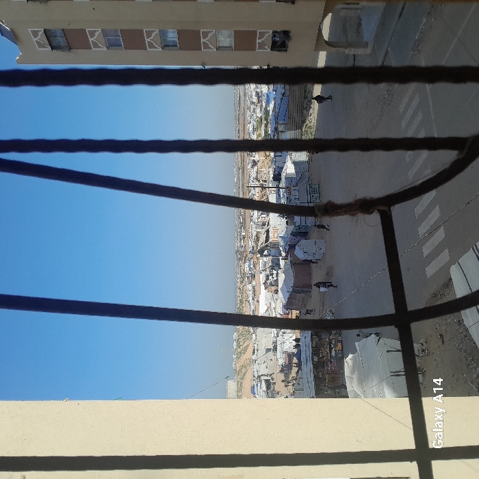 A partially obscured view from a window looking out over a settlement. Tents and low buildings spread across a dusty landscape under a clear blue sky. A pedestrian crossing marks a road adjacent to the settlement. The scene is framed by the window's bars. A multi-story building appears at the top.