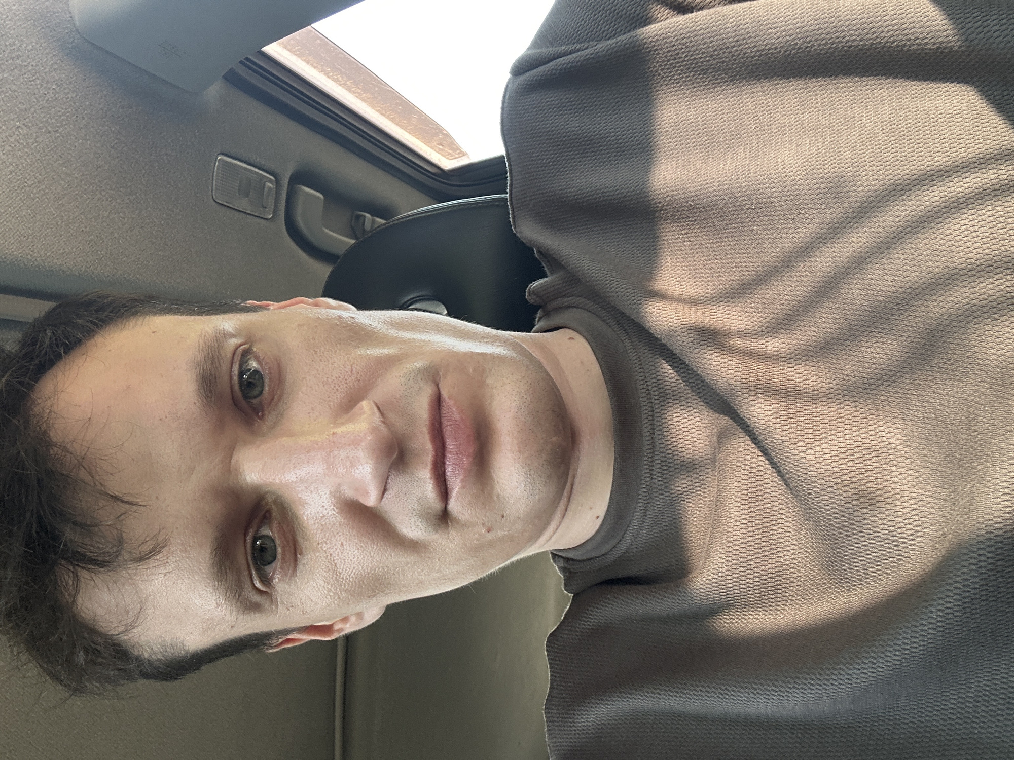 A close-up, high-angle, selfie-style shot shows a fair-skinned man with short, dark hair, taken inside a vehicle. The man faces the camera, his eyes looking forward, and his lips are slightly parted. He wears a dark grey, waffle-knit long-sleeved shirt. Sunlight streams in from the right, casting shadows across his face and shirt. The background shows the interior of a car, including the headliner and a portion of a dark car seat. The man's facial features are prominent, with a relatively narrow face and sharp jawline. He appears to be in his late 20s to early 30s. The image does not contain any information or visual cues that would allow for the determination of its specific location within the city of Makiv, Ukraine.