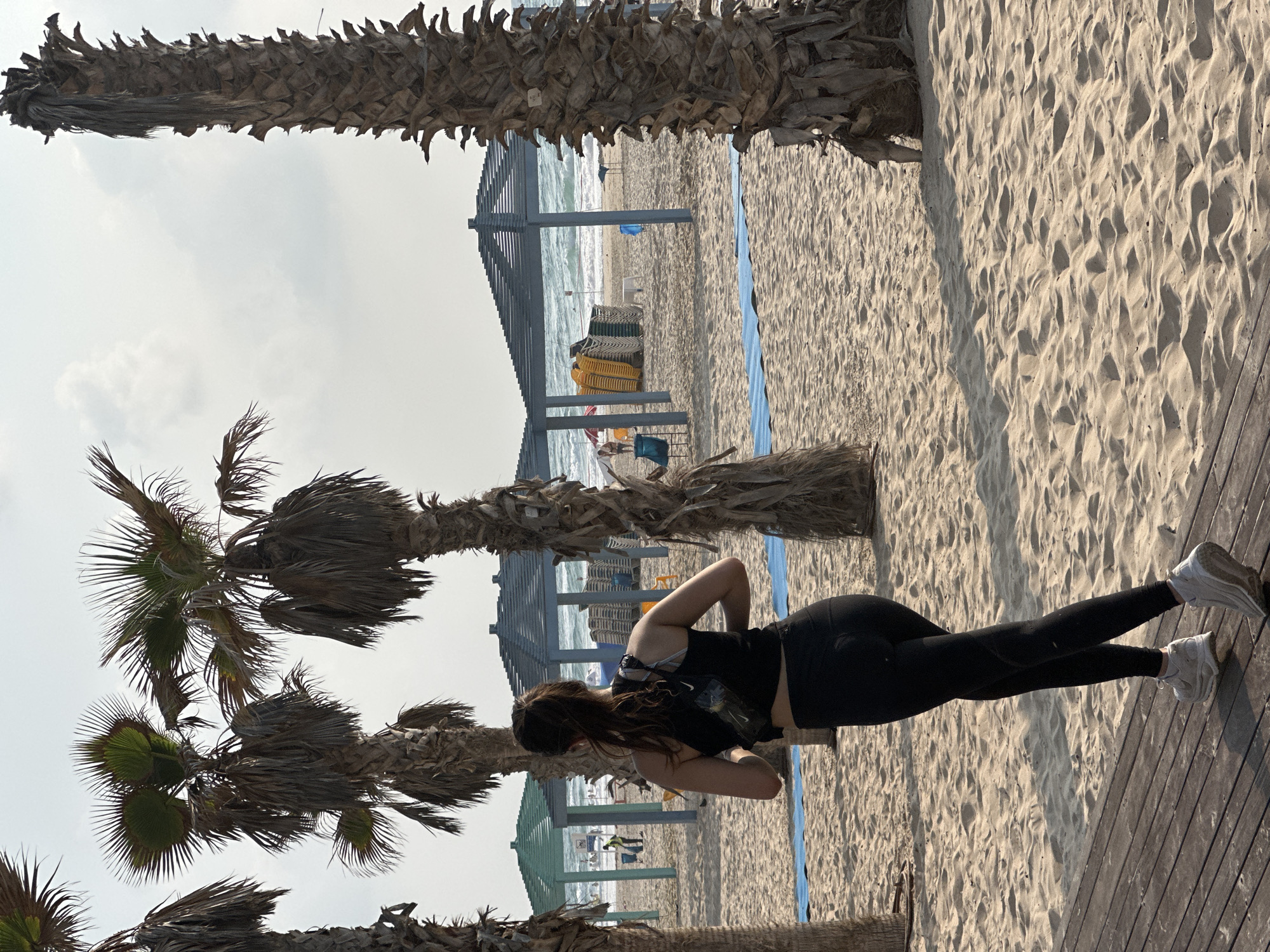 The image depicts a woman with dark hair in a ponytail, wearing a black tank top and black leggings, as she walks on a wooden walkway towards a sandy beach in Bat Yam, Israel. She is facing away from the camera, her right arm bent at the elbow. She has a black waist bag or fanny pack. Behind her, the sandy beach is lined with palm trees and beach umbrellas. In the background, the sea with waves is visible, along with beach chairs stacked under the umbrellas. The sky is overcast with some clouds. The overall scene suggests a recreational setting on a beach, possibly during the day.
