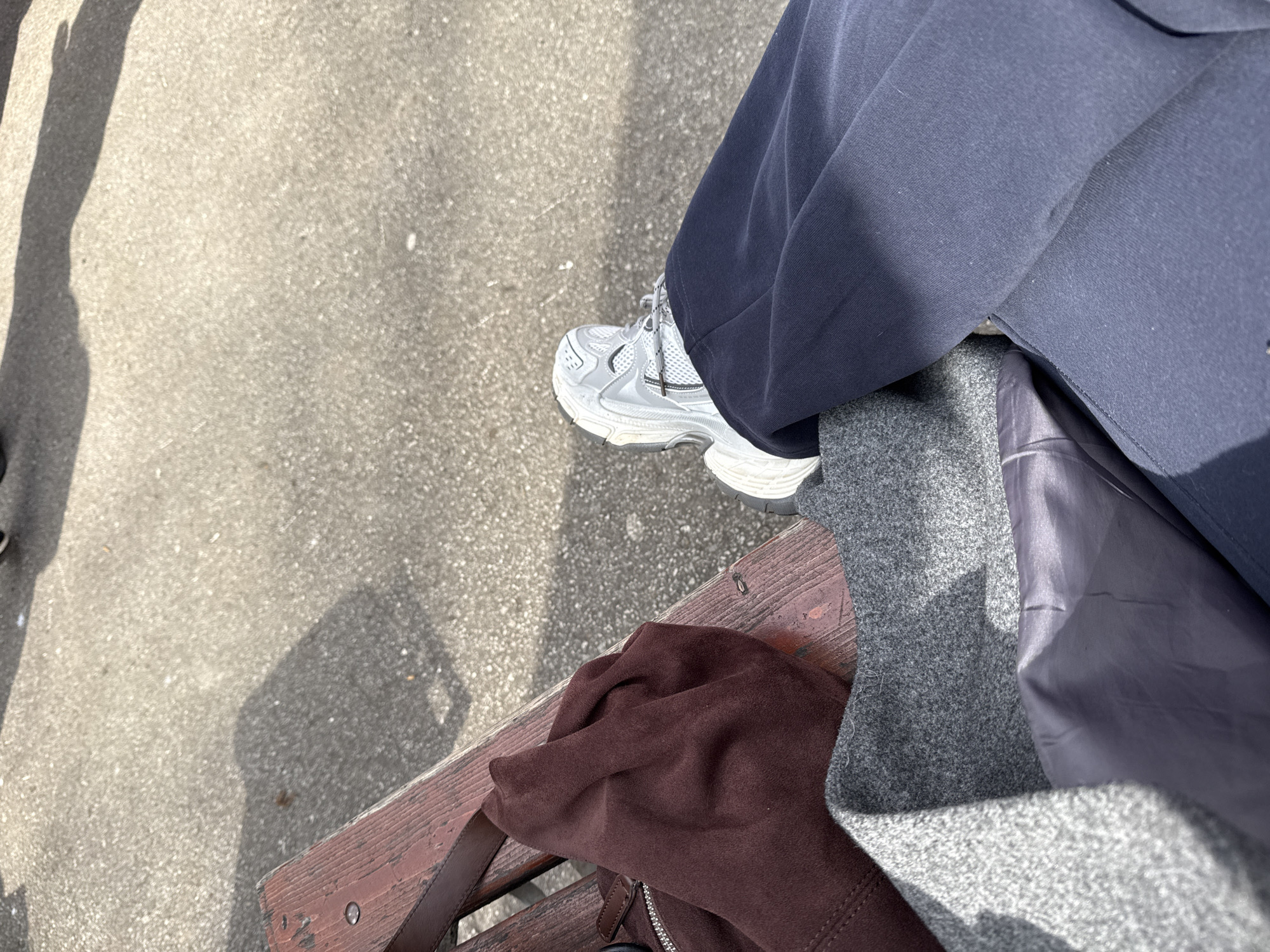 A photograph from an elevated perspective shows a person seated on a weathered wooden bench, likely in an urban outdoor space. The bench is made of reddish-brown slats with worn edges. The person is wearing loose-fitting, dark blue trousers and a gray and light gray athletic-style sneaker. Resting on the bench beside the person's leg is a brown suede or leather bag with a visible zipper and strap. A gray wool-like coat is also visible, with a darker gray or blue lining. The background is a concrete or paved surface with shadows cast by unseen objects. The details lack specificity needed to verify the location in the city of Perm, Russia.