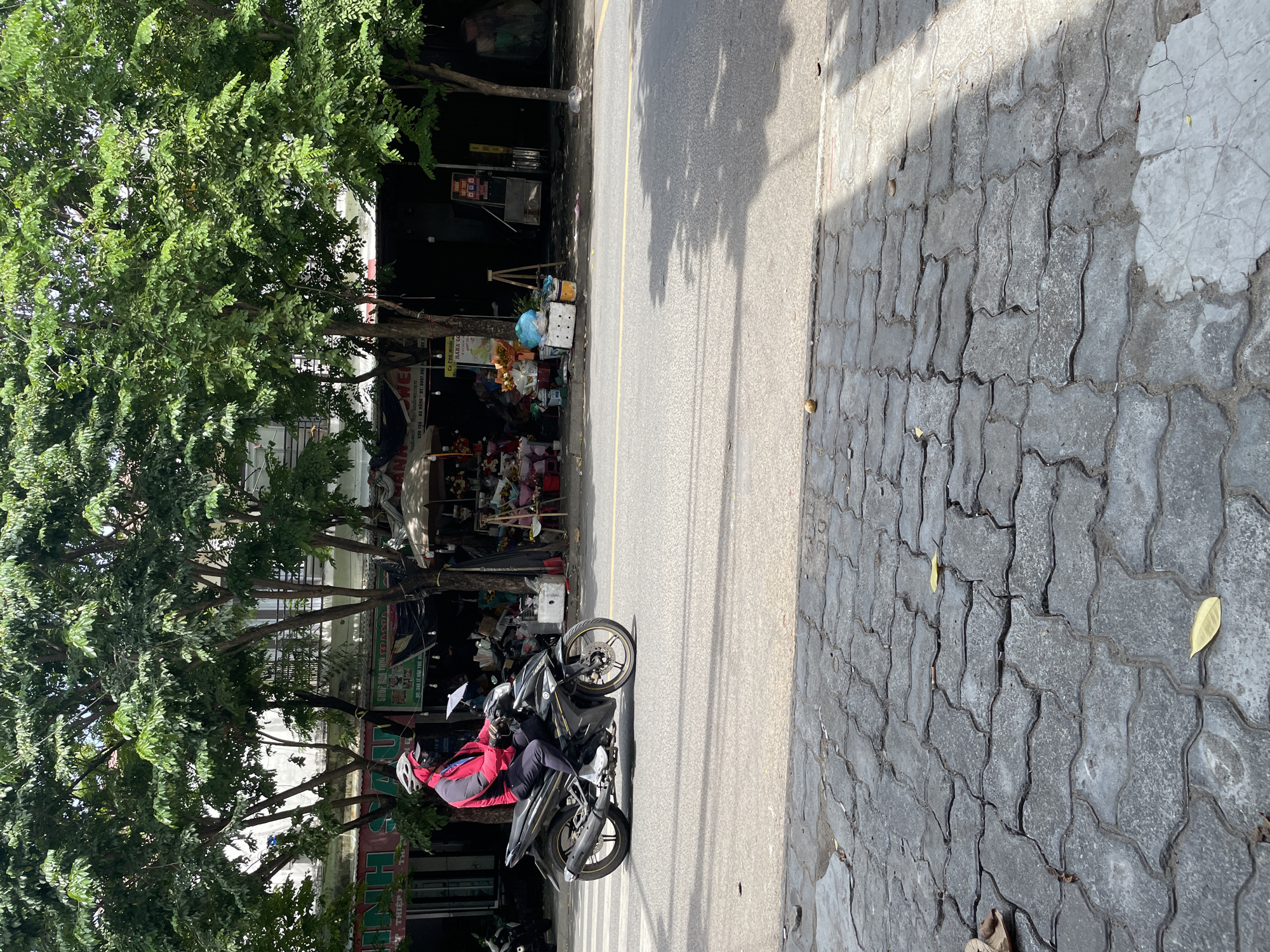 A person on a black motorcycle rides on a street in Đà Nẵng, Vietnam. The person is wearing a red and grey jacket, black pants, and a white helmet. The motorcycle is a sport bike with gold accents. The street is paved with asphalt and has a yellow line dividing the lanes. On the right side of the street, there are trees and shops. The shops are decorated with flowers and other items. The pavement on the left side of the street is made of interlocking concrete blocks. The sky is cloudy.