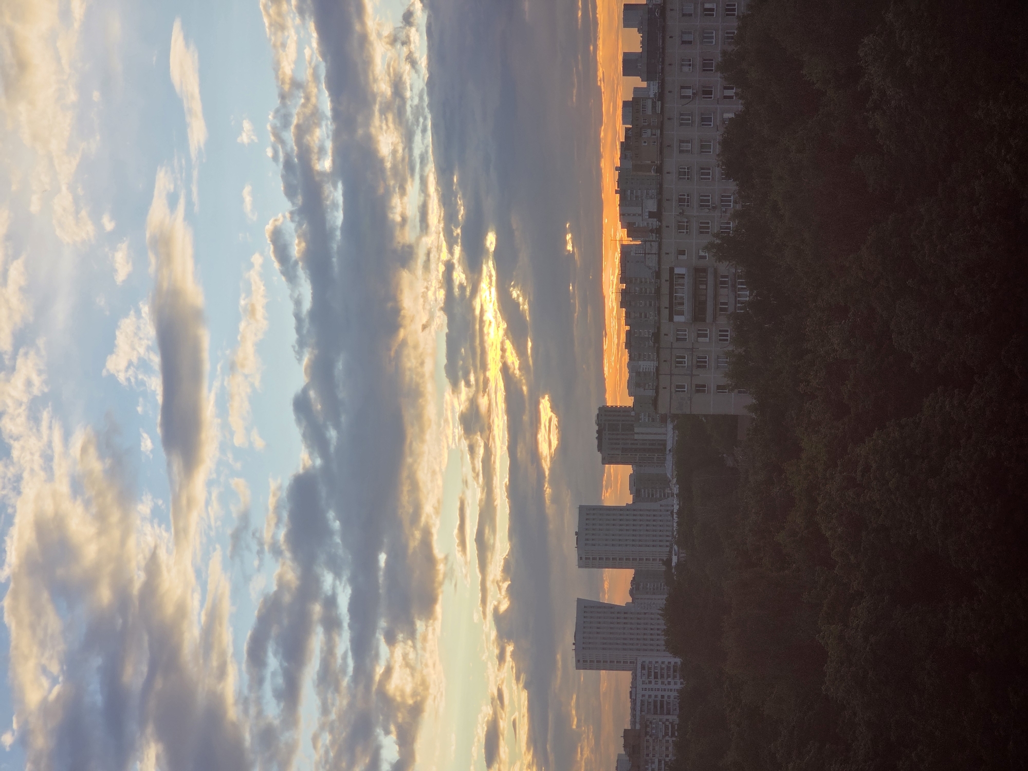 The image depicts a city skyline at dusk, likely in Moscow, Russia, given the architectural style of the buildings and the prevalent cloud formations. The sky is a dramatic canvas of oranges, yellows, and blues, with heavy, textured clouds catching the last rays of the setting sun. Below the sky, a dense canopy of dark green trees dominates the foreground, obscuring much of the lower portion of the cityscape.
Rising above the trees are several multi-story buildings, including a cluster of modern, tall white buildings with numerous windows. To their right, and closer to the foreground, is a long, blocky, grey residential building characteristic of Soviet-era architecture, featuring repetitive window patterns and balconies. In the distance, more buildings are visible, their shapes softened by the fading light and atmospheric haze. The overall impression is one of an urban landscape at the end of the day, with nature encroaching on the built environment. There are no people or discernible actions visible in the frame.
