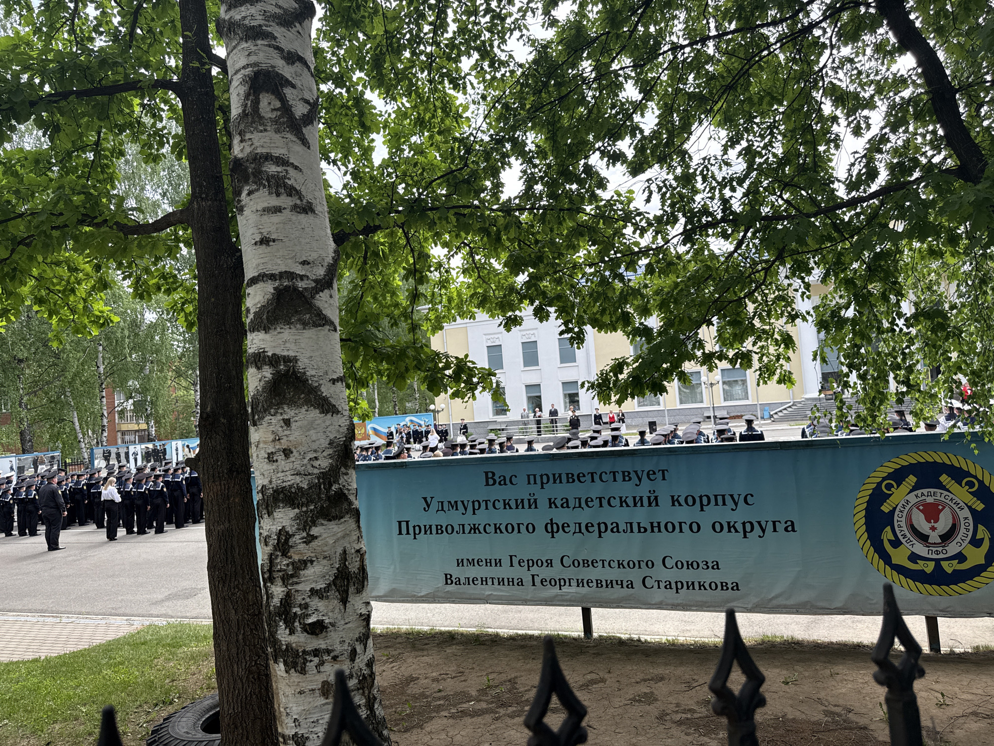 The image shows an outdoor event likely in Votkinsk, Russia, judging by the text on the banner. The banner reads "Welcomes You: Udmurt Cadet Corps of the Volga Federal District, named after Hero of the Soviet Union Valentin Georgievich Starikov." A group of people in dark uniforms and hats are lined up in the background, possibly cadets participating in a ceremony. The scene is partially obscured by trees in the foreground, including a distinctive birch tree. A black fence is visible in the immediate foreground. A building with white walls and yellow accents is in the background behind the lined-up group. The sky appears overcast.