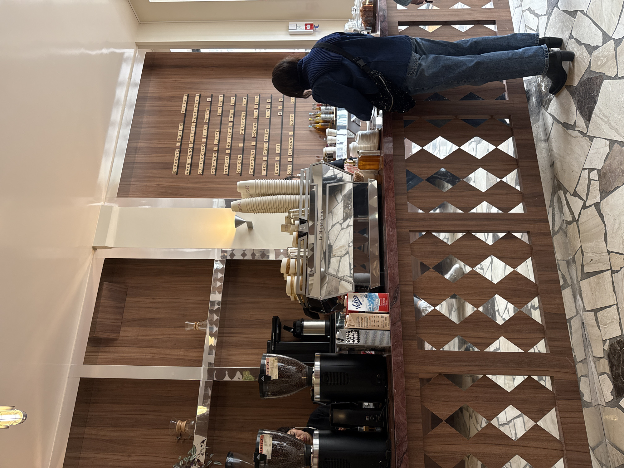 A person with dark hair, wearing a dark blue jacket and blue jeans, stands at a wooden counter in what appears to be a cafe. On the wall behind the counter, a menu lists various coffee and beverage options along with prices, including "Espresso," "Americano," and "Latte." A modern espresso machine sits on the counter, next to stacks of paper cups and containers. Below the counter, a coffee grinder is visible. A shelf with coffee beans is also present. The countertop is decorated with a geometric pattern. The floor alongside the counter has a mosaic tile design. Based on the decor and product names, the cafe is likely located in Paris, France.