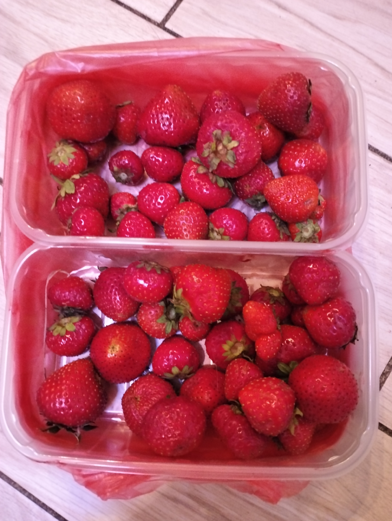 Two clear plastic containers are stacked atop one another, both filled with fresh, red strawberries. The strawberries vary in size, from small to large. The containers rest on a light-colored surface with faint linear patterns. The scene is indoors. No people are visible. There is no indication of Krykhivtsi, Ukraine.