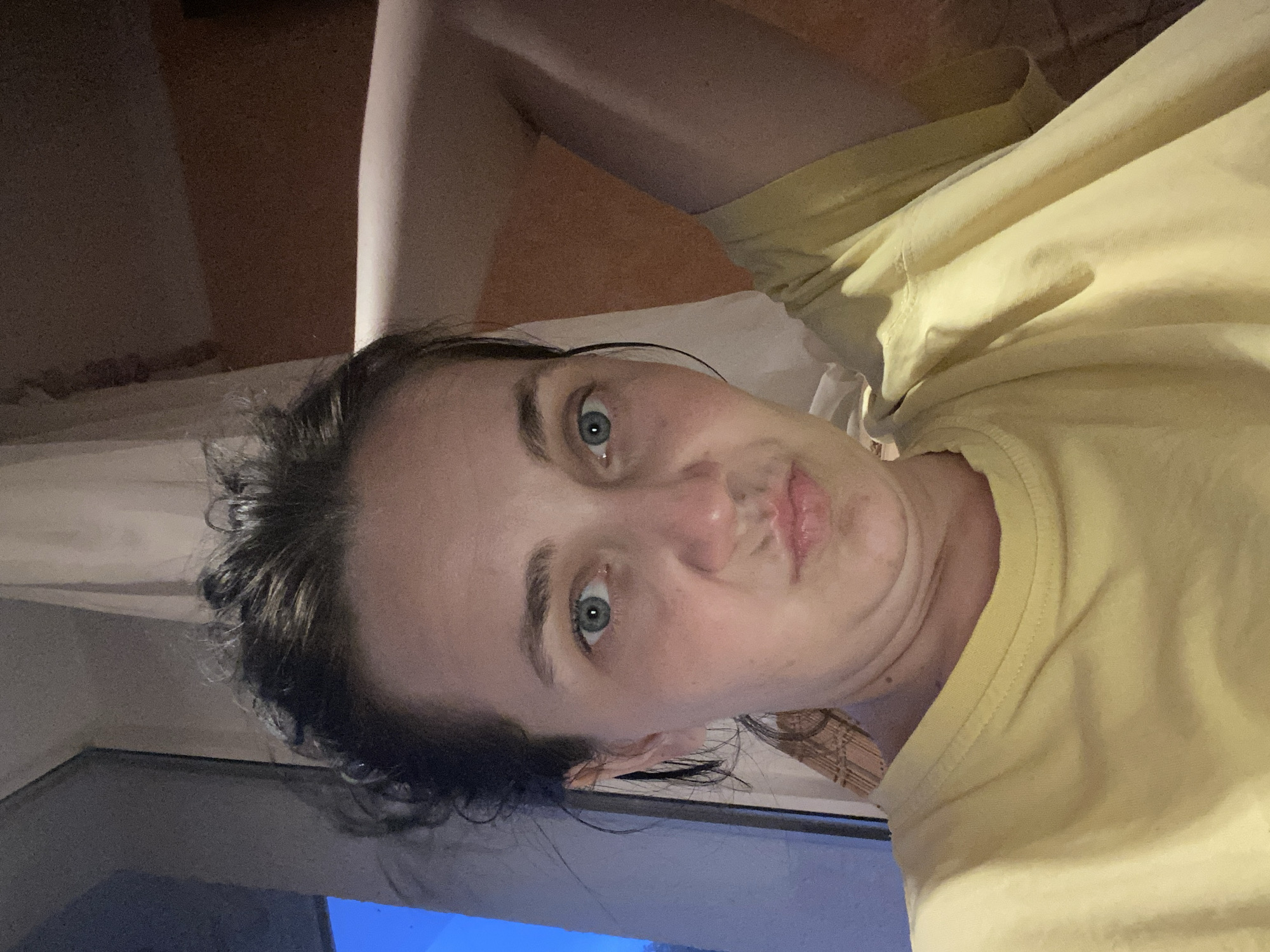 An over-the-shoulder, medium shot captures a woman from the chest up as she poses with her left arm behind her head and her right arm bent and resting on her chest. The woman is facing the camera with her head tilted slightly down and to her left. She has light skin, dark brown hair pulled up into a messy bun, and blue eyes. Her eyebrows are dark and arched. She has a neutral expression with her lips pursed. She is wearing a yellow t-shirt. The background shows a window on the left with a dark blue light, and a curtain to the right of the window. The right side of the background is a textured wall in a peachy-orange color. The lighting appears to be artificial and coming from the front and right of the woman.