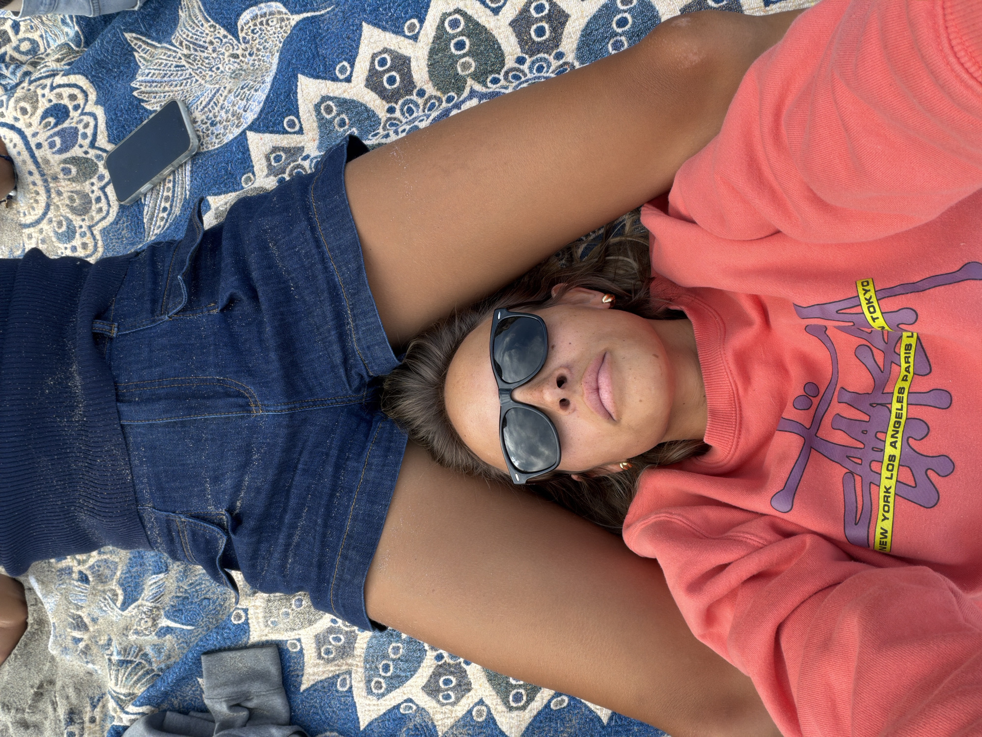 A woman is lying on a patterned blue and white blanket at a beach, with her head resting on the thighs of another person wearing denim shorts. She is wearing sunglasses and a pink sweatshirt with text including "NEW YORK LOS ANGELES PARIS LONDON TOKYO." A mobile phone is near the denim shorts. The scene suggests a casual, relaxed setting. I have no information to prove the location as Malibu, United States of America.