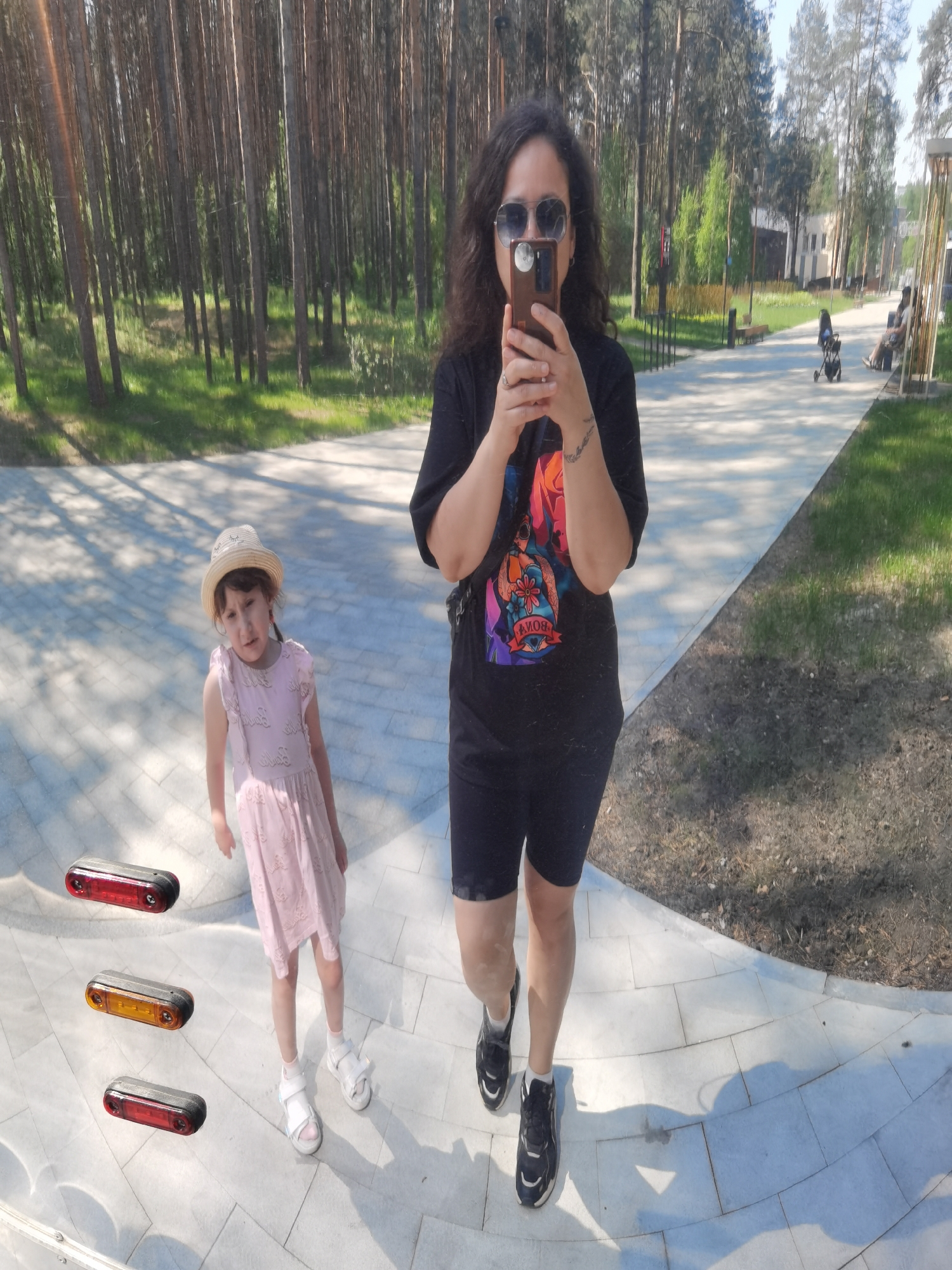 In a well-lit outdoor scene, a woman with curly hair and sunglasses is holding a smartphone up to take a picture. She is wearing a black t-shirt with a colorful graphic design and black shorts, along with black and white sneakers. A young girl, wearing a light pink dress and a straw hat, stands to the left of the woman. Three rectangular lights with red and orange lenses are digitally added near the girl. They are on a sidewalk, which appears to be part of a park or public area. In the background are tall trees, indicating a wooded or park-like environment, and other people seated on benches. This is likely a recreation area or park, possibly located in the city of Perm, Russia.