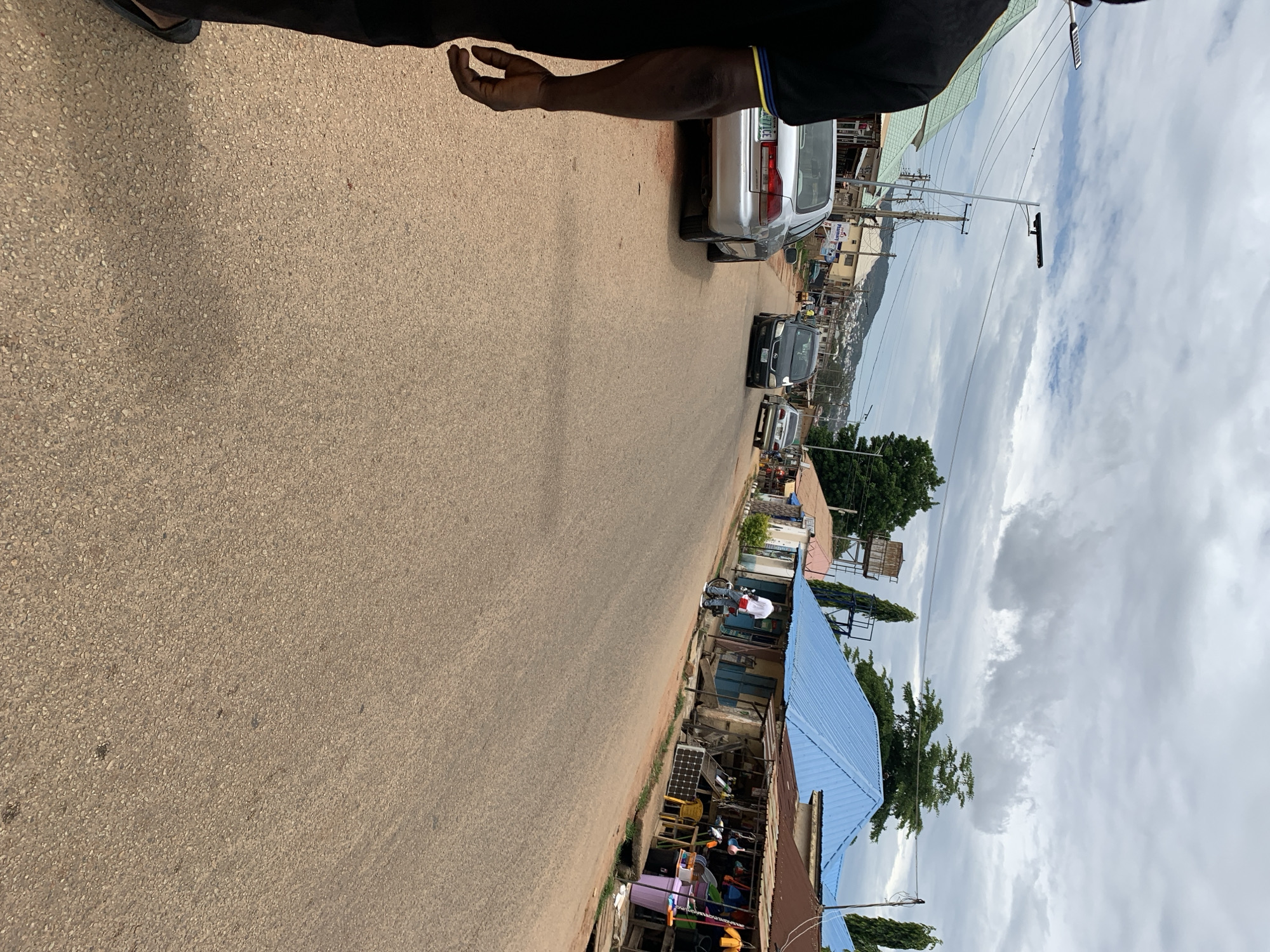 This image depicts a wide-angle street-level view of a thoroughfare in New Stadium, Nigeria, under an overcast sky filled with stratocumulus clouds. The perspective is from behind a person standing on the left side of the road, looking down the street.
The road surface is a dry, light brown, coarse asphalt or gravel, extending from the immediate foreground into the distant background, appearing relatively flat with a slight curve to the left.
In the immediate foreground, the back of a person is visible on the left. This individual wears a black short-sleeved top with a narrow blue and yellow stripe on the collar and dark trousers. A dark flip-flop or sandal is visible on the left foot, and the person's right forearm and hand are partially visible.
On the left side of the road, a silver four-door sedan (appearing to be a Honda Accord) is parked facing away from the camera. Its license plate, "FG 771CE," is partially legible. Beyond this car, a row of single-story buildings with green corrugated metal roofs is visible, interspersed with wooden utility poles carrying numerous electrical wires. One building displays a sign with the word "Pharmacy." Two tall poles with solar-powered streetlights are also present on this side of the road.
On the right side of the road, a dark-colored four-door sedan is approaching the camera in the mid-ground, followed by a lighter-colored sedan further in the distance. Adjacent to the road, a person in a white t-shirt and blue jeans is standing next to a white motorcycle or scooter, parked on the unpaved shoulder. Beyond this, a series of informal market stalls and single-story buildings line the street. One building features a distinctive bright blue corrugated metal roof. The market stalls have darker, rusted corrugated metal canopies and display various household goods, plastic buckets, brooms, and other items. A yellow plastic chair and a yellow-framed solar panel are positioned outside one of these stalls. Several indistinct figures are visible within the market area, some seated on yellow plastic chairs.
In the mid-ground, behind the buildings on the right, a rusty metal water tank supported by a tall metal frame is prominent. Various green trees are scattered throughout the scene, and a range of green hills is visible on the horizon in the background.