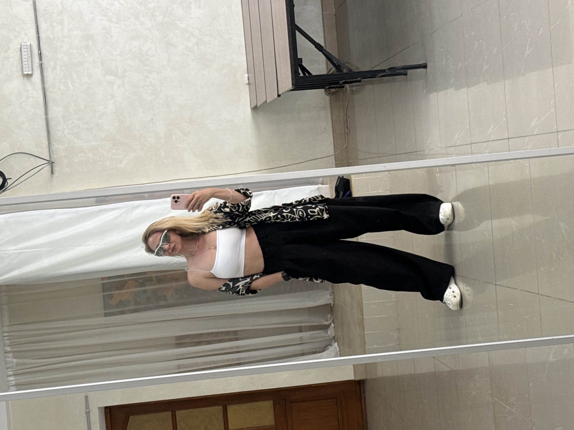 The image is a reflection of a person taking a selfie in what appears to be a bathroom or dressing room. The person is wearing a white top, black wide-leg pants, a patterned jacket, and white shoes (possibly Crocs). They are holding a pink phone up to capture the image. The background includes a white curtain, a dark wooden door, and tiled walls. The exact city or location cannot be determined from the given image.