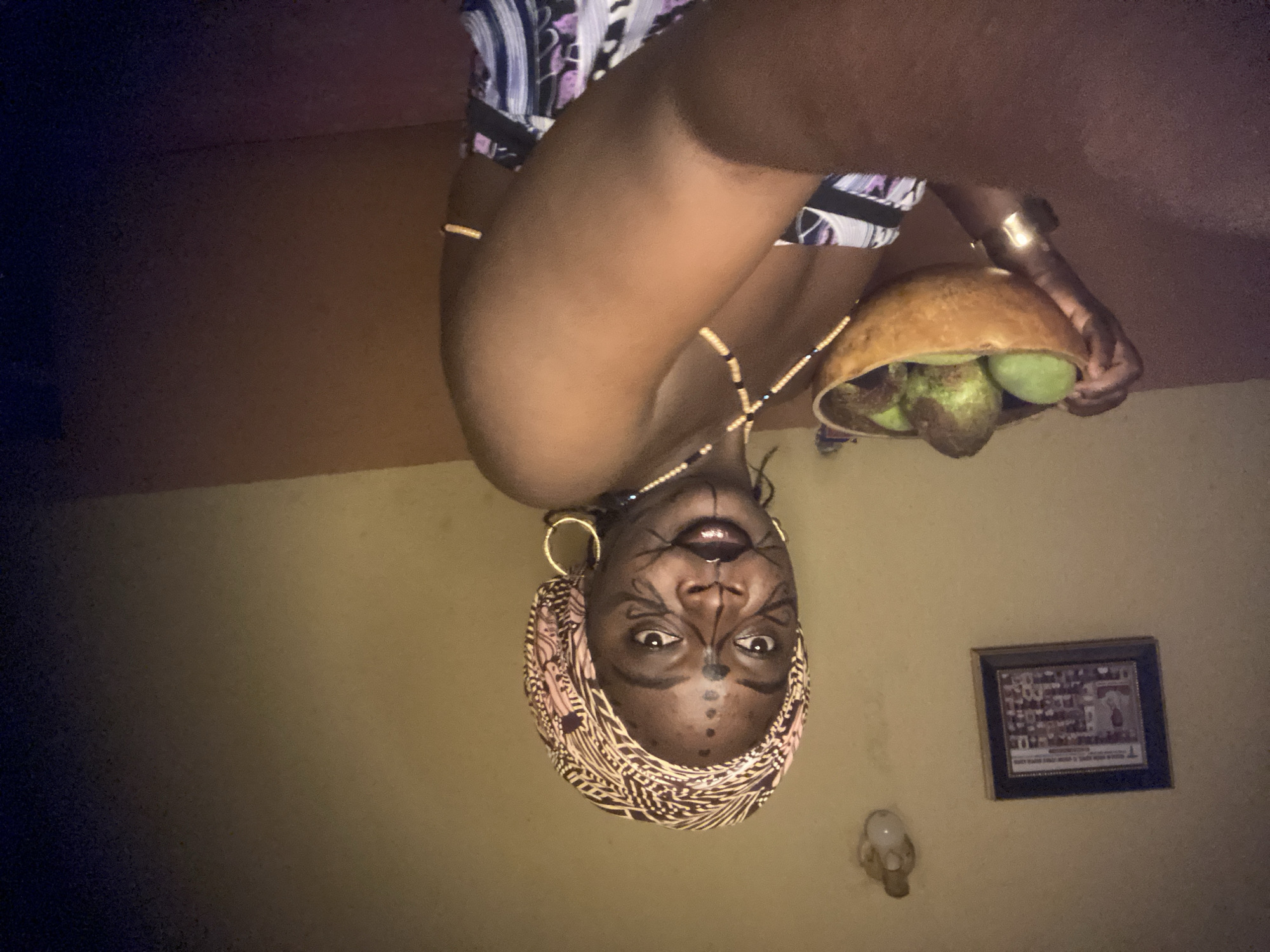 A woman with tribal face paint and a patterned headscarf is holding a gourd filled with green fruits, possibly avocados. She is wearing gold hoop earrings and a necklace with dark beads. The background shows a light yellow wall, a framed picture, and a wall sconce. The image appears to have been taken indoors. The location is Maigizo, Nigeria.