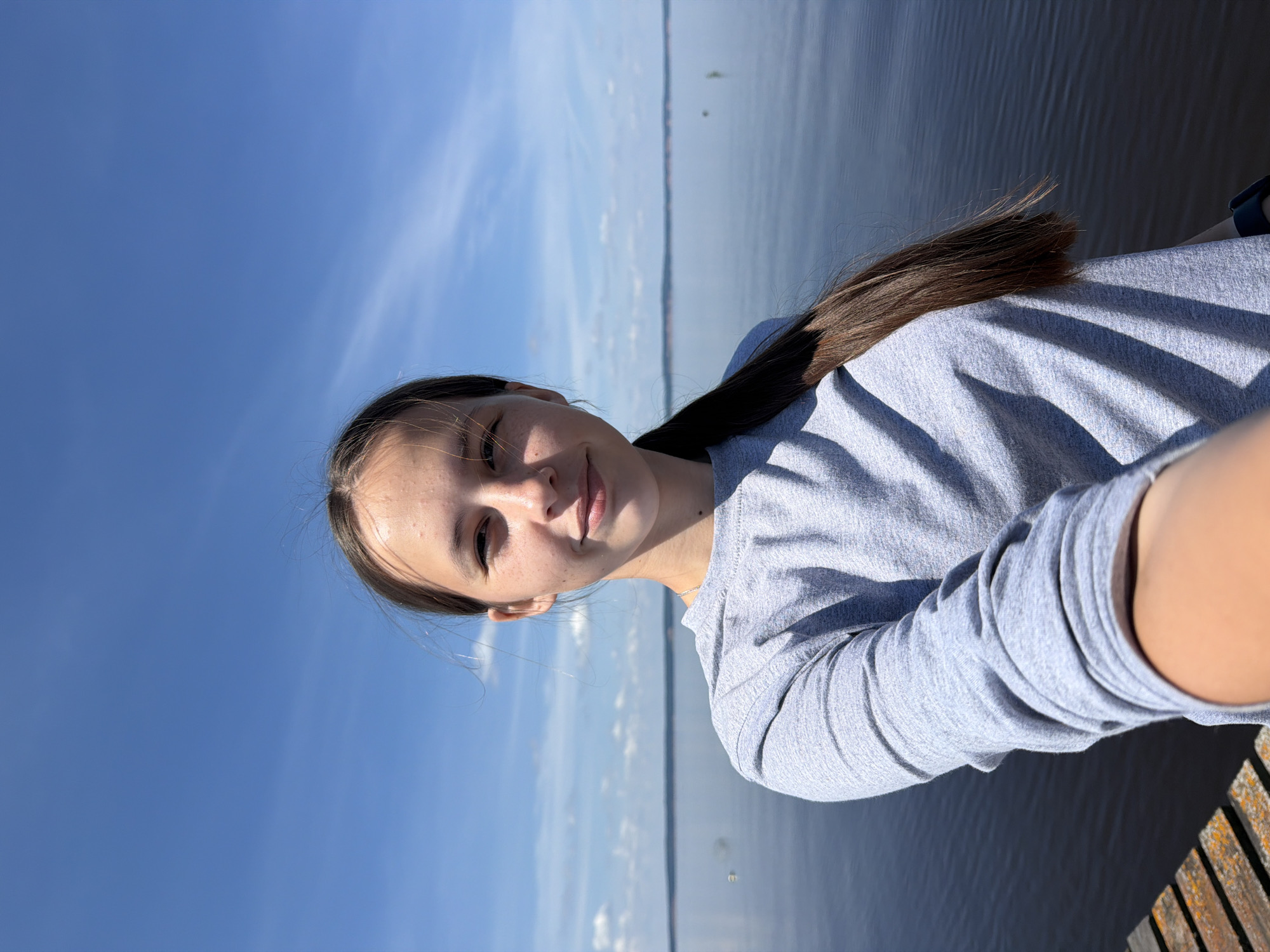 A young woman with long brown hair pulled into a ponytail takes a selfie on a clear, sunny day. She is wearing a light grey long-sleeved shirt and is standing on a wooden pier, with a large body of water behind her. The water stretches to the horizon, where it meets a distant treeline under a bright blue sky dotted with wispy white clouds. The pier in the foreground is made of weathered wooden planks. The scene is consistent with a location in or near Stepanovo, Russia, given the typical landscape of the region.