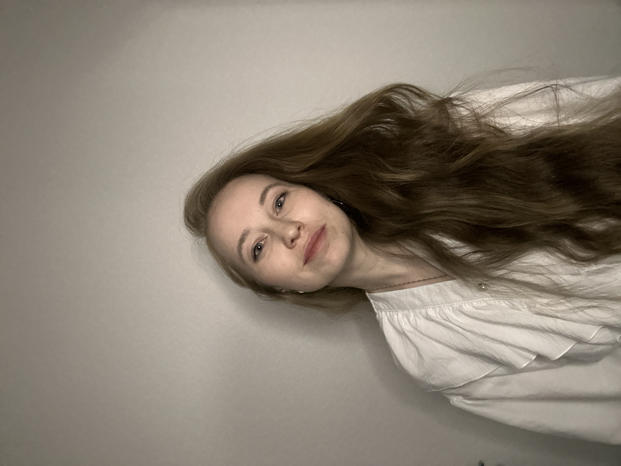 A woman with long brown hair is facing the camera at an angle. She is wearing a white, ruffled blouse. The background is a plain, light gray wall. The location cannot be determined from the given image.