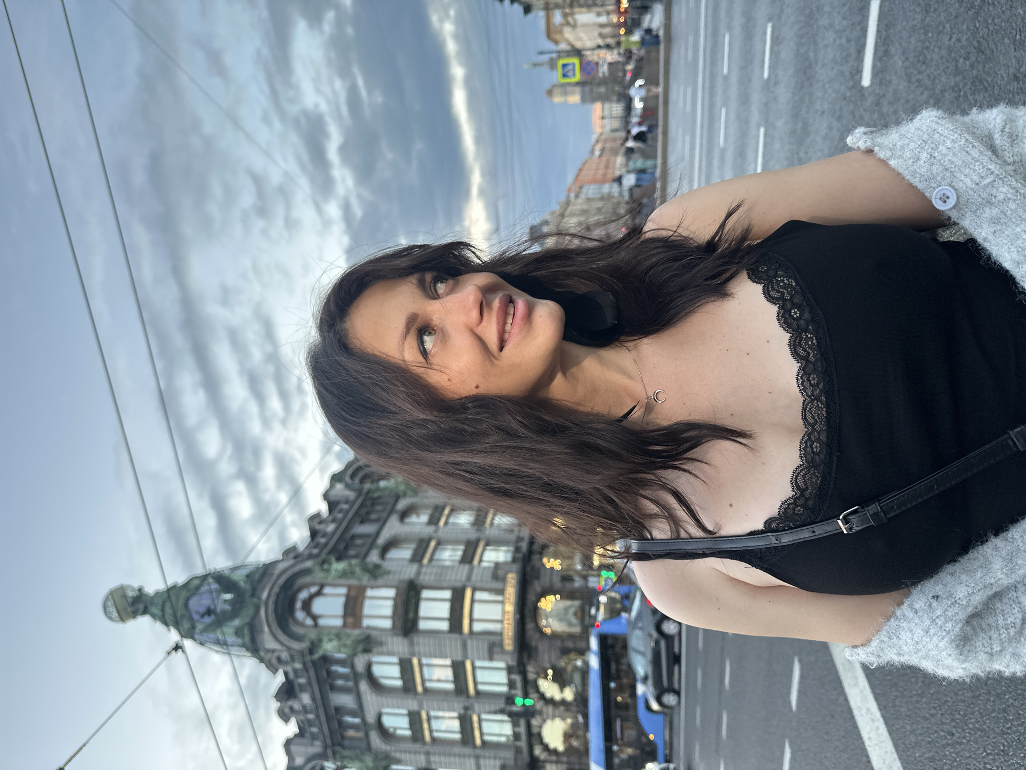 A woman with dark hair, wearing a black camisole with lace trim and a grey cardigan draped over her shoulders, stands on a street in Saint Petersburg, Russia. She is looking to her right with a slight smile. Behind her, a large building with a distinctive green dome topped by a globe is visible, along with a blue bus and other city traffic. The sky is overcast with grey clouds. The scene appears to be set during the evening.