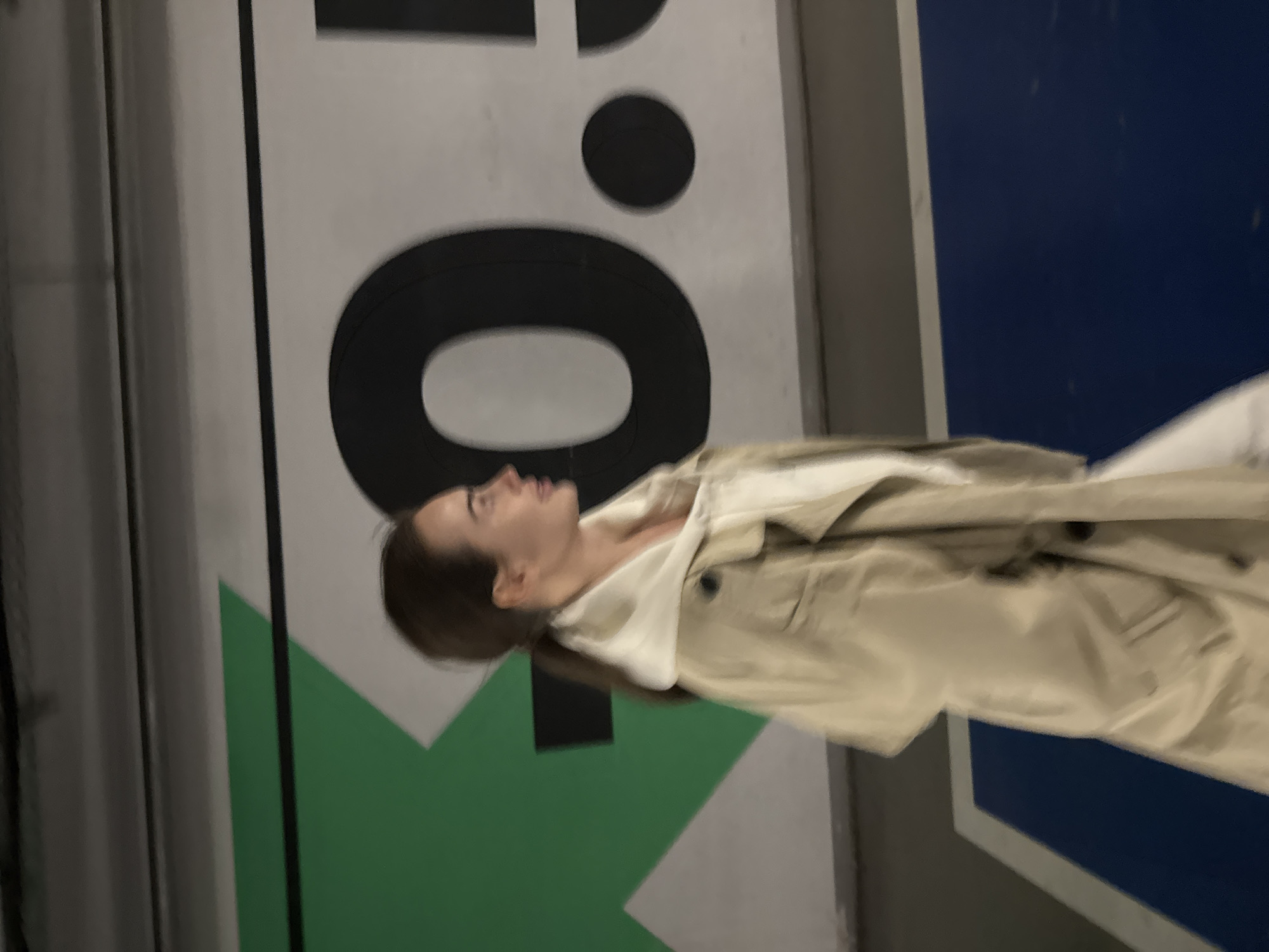 The image depicts a young woman in what appears to be an indoor or covered urban setting, possibly in Warsaw, Poland. She is positioned centrally, viewed in profile, facing towards the right and looking upwards with a neutral, contemplative expression. Her dark hair is styled back, and she is dressed in a light beige or tan trench coat worn over a lighter-colored inner layer.
The background is dominated by a large graphic sign or wall. Prominently featured on a white surface is a large, bold, black sans-serif letter "O", accompanied by a smaller black circle located above and slightly to its right. Below and to the left of these characters, a sharp, green geometric shape, resembling a triangle, is visible. To the right of the woman, the background transitions into a solid dark blue section. On the far left, a vertical grey column or divider frames the scene. The lighting is artificial and muted, suggesting an indoor environment or evening conditions. The woman's gaze directed upwards implies she is observing something on or above the sign in this modern public space.