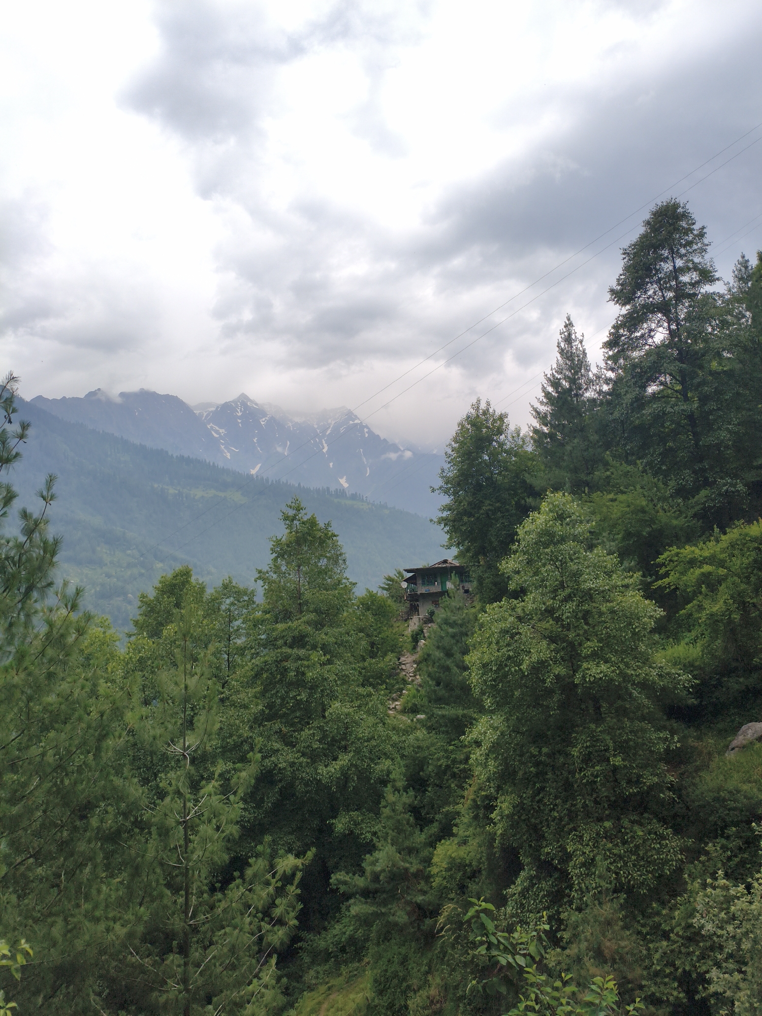 In the city of Vashisht, India, a picturesque view unfolds. The image captures a majestic mountain range in the distance, its peaks dusted with snow, partially obscured by a cloudy sky. In the mid-ground, a cluster of trees forms a dense forest, with a small, rustic house nestled on a slope amidst the foliage. The foreground is dominated by lush green trees, their branches reaching towards the sky, creating a natural frame for the mountainous backdrop. The overall scene evokes a sense of serene natural beauty and tranquility.