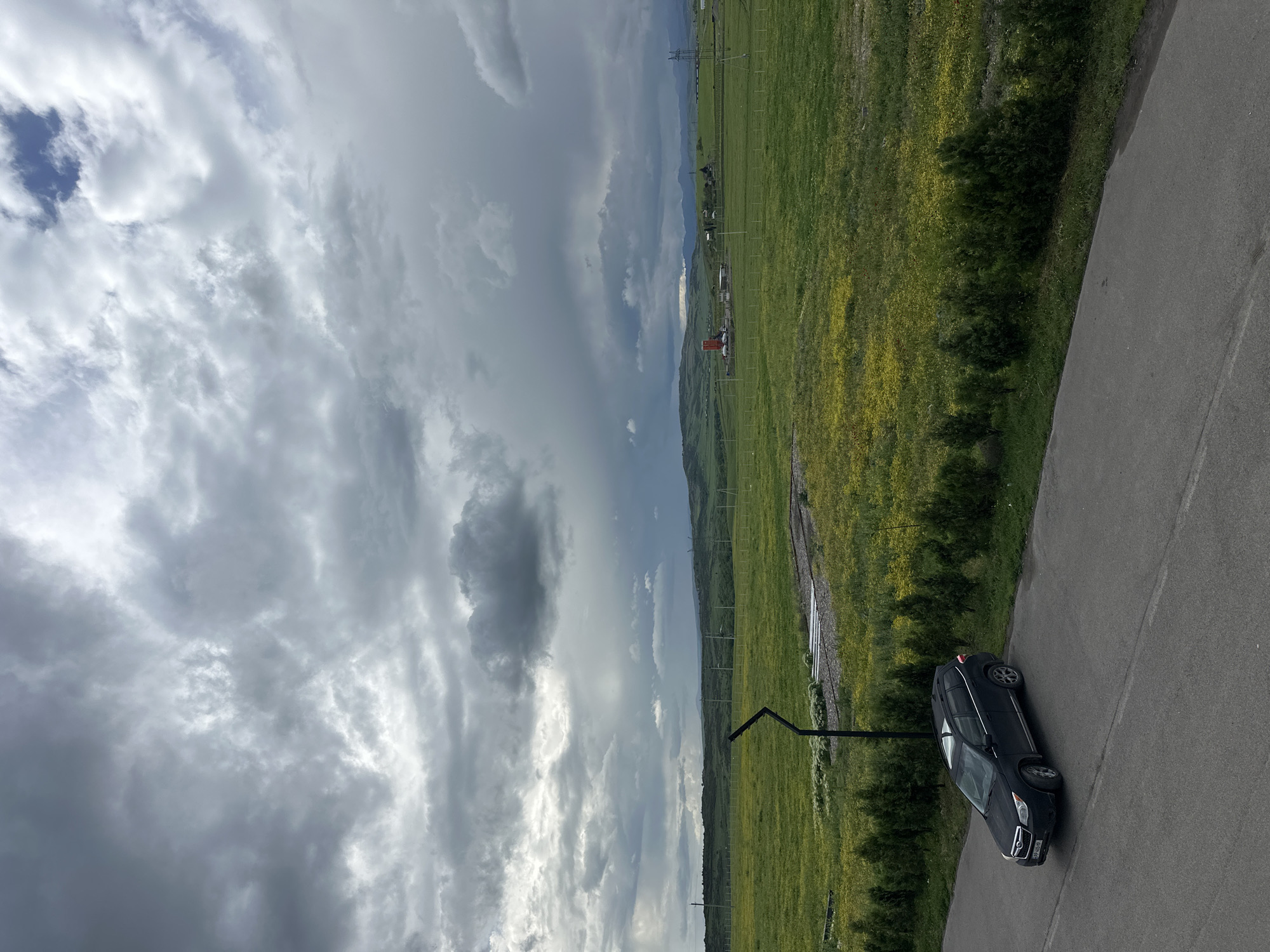 A black SUV is parked on an asphalt road adjacent to a green field. The road stretches into the distance, and the field is bordered by trees and vegetation. In the background, a landscape of rolling hills and mountains can be seen under a cloudy sky. The location is in the city of Marneuli, Georgia.