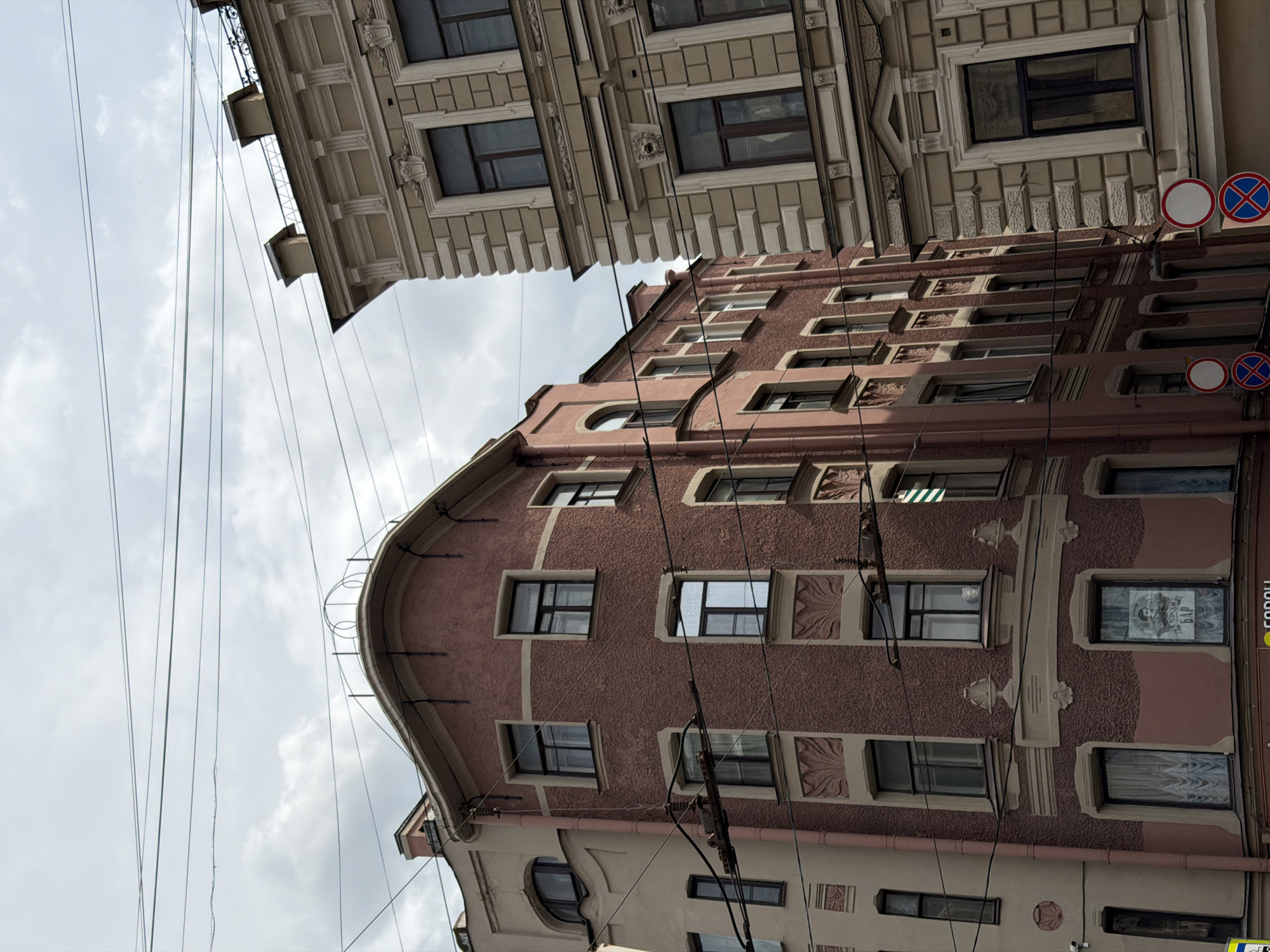This is a photograph of two buildings in Saint Petersburg, Russia, with overhead power lines visible against a cloudy sky. The building on the left is a corner building with a rounded facade, painted a muted pinkish-tan and a darker pinkish-brown. It features multiple windows of various sizes, some with decorative trim. The building on the right is an older structure, lighter in color, with ornate architectural details around its windows and a prominent cornice. The lower parts of the buildings are not fully visible, but some street signs and the upper portion of what appears to be a bus or tram can be seen at the very bottom edge of the frame. There are no people clearly visible in the image. The perspective is from a low angle, looking up at the buildings.