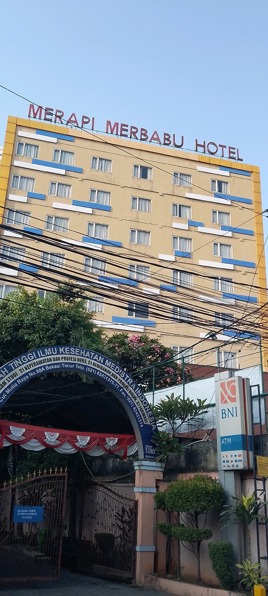 This image shows the MERAPI MERBABU HOTEL building in Bekasi, Indonesia. The hotel has a cream-colored facade with blue and white horizontal accents. There are many power lines in front of the building. Below the hotel, there is an arched entrance with text that reads "AKADEMI TINGGI ILMU KESEHATAN MEDISTRA INDONESIA". The entrance is decorated with red and white fabric banners. A gate is visible to the left of the entrance. To the right of the entrance is a BNI ATM sign. The environment appears to be an urban area with trees and some greenery.