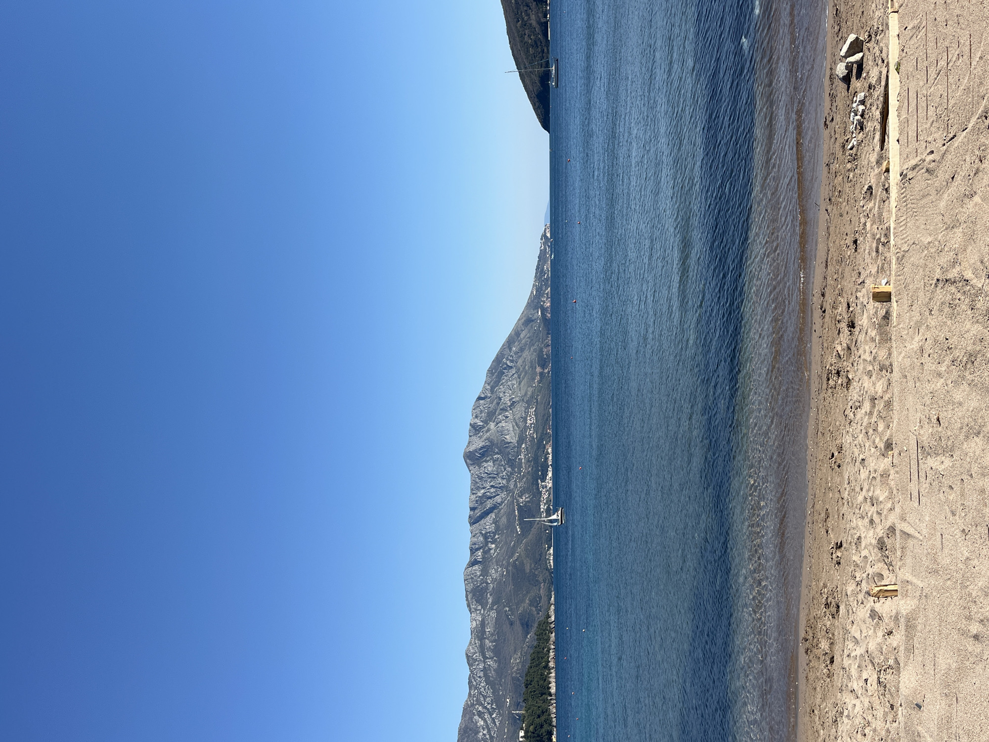 The image shows a scenic view of a beach in Budva, Montenegro. The clear blue sky contrasts with the dark blue water, where a single sailboat can be seen. A tall mountain range lines the opposite side of the water. The beach itself is light-colored sand with wooden planks on the edge. No people are visible in the image.