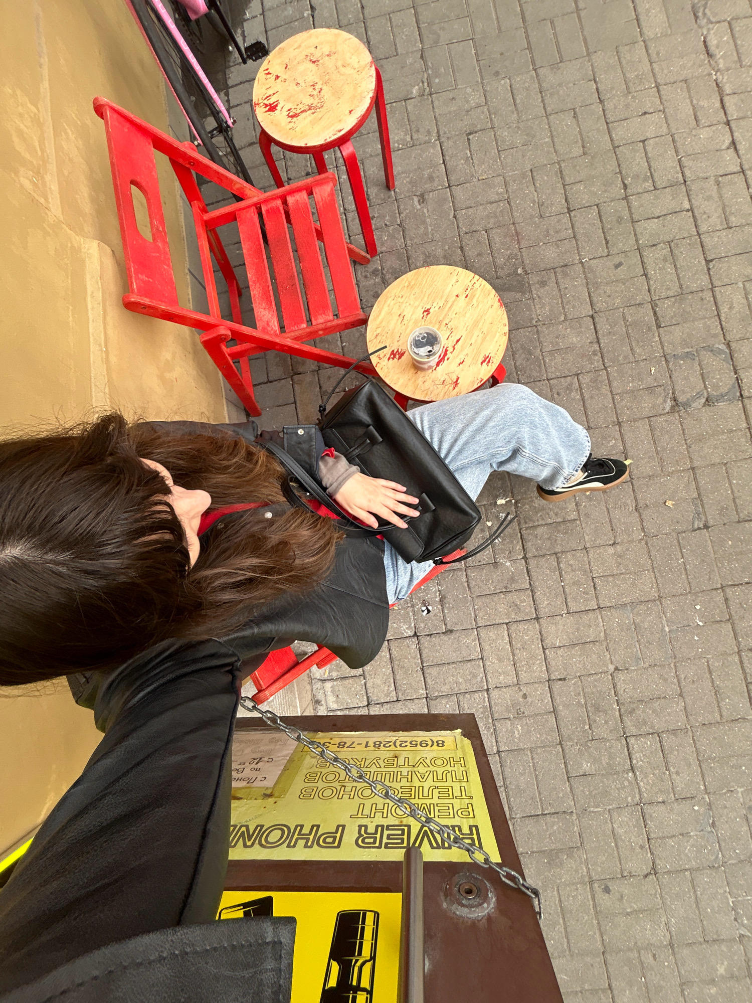 A person sits at a small, circular wooden table outside a building. The person has long, dark brown hair and is wearing light blue jeans and black sneakers. They are holding a black purse. A red chair is positioned behind them, and another red stool is to their left. The ground is paved with gray bricks. A yellow sign is visible on the ground, featuring the words "RIVER PHON" in large letters, along with some Russian text and a phone number. The sign also shows an image of a microphone. The overall scene appears to be an outdoor cafe or seating area in a city. The sign with Russian text indicates the location is likely Saint Petersburg, Russia.