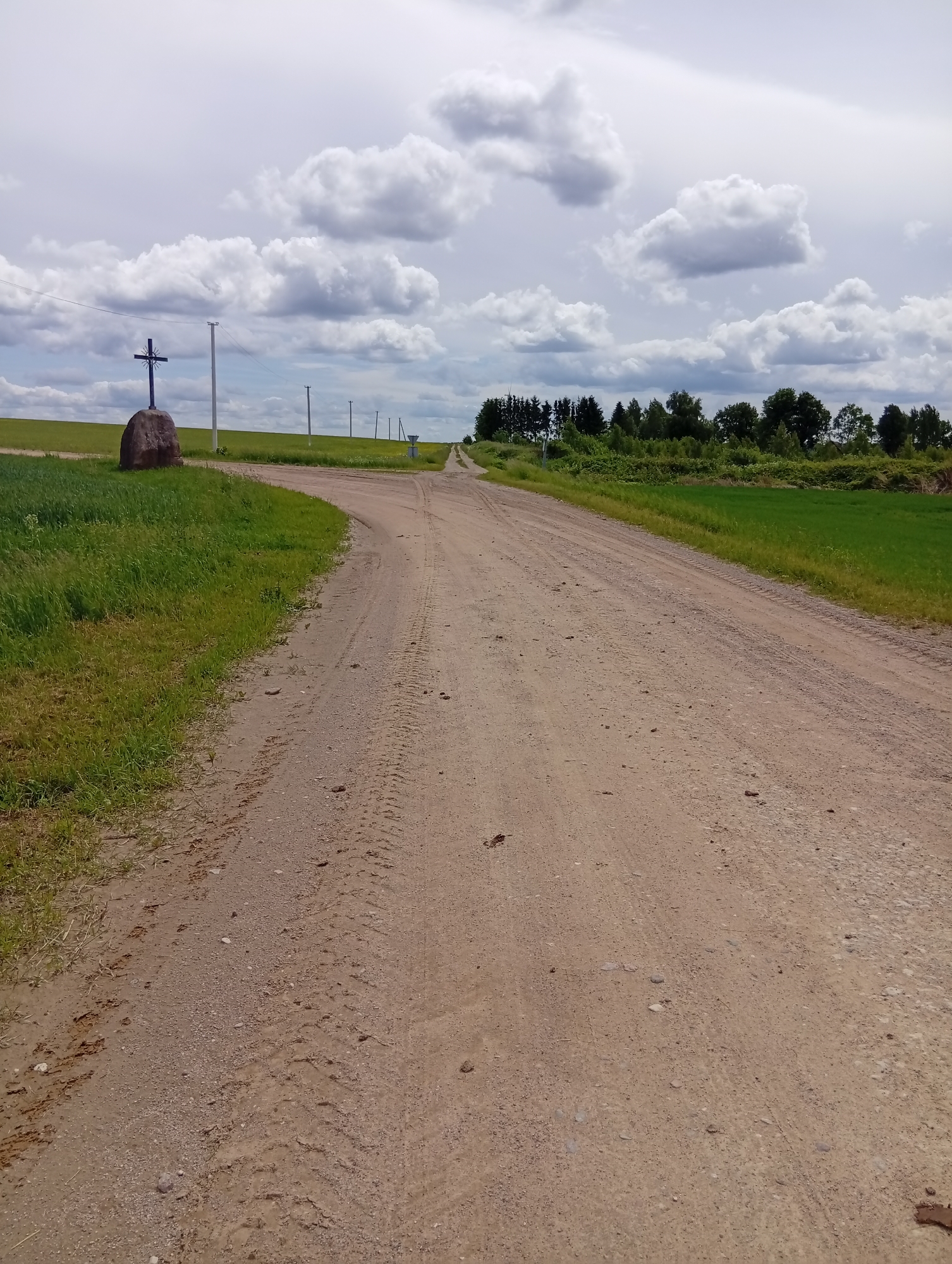 A dirt road curves to the left in the foreground, leading towards a treeline in the distance. On the left side of the road, a grassy field slopes upwards, and a large, dark stone stands with a metal cross affixed to its top. In the background, a line of utility poles stretches across the horizon, with a cluster of trees visible further back. The sky is filled with dramatic, grey and white clouds. The scene is likely located in Krikliai, Lithuania, given the presence of a roadside cross, a common landmark in rural Lithuanian landscapes.