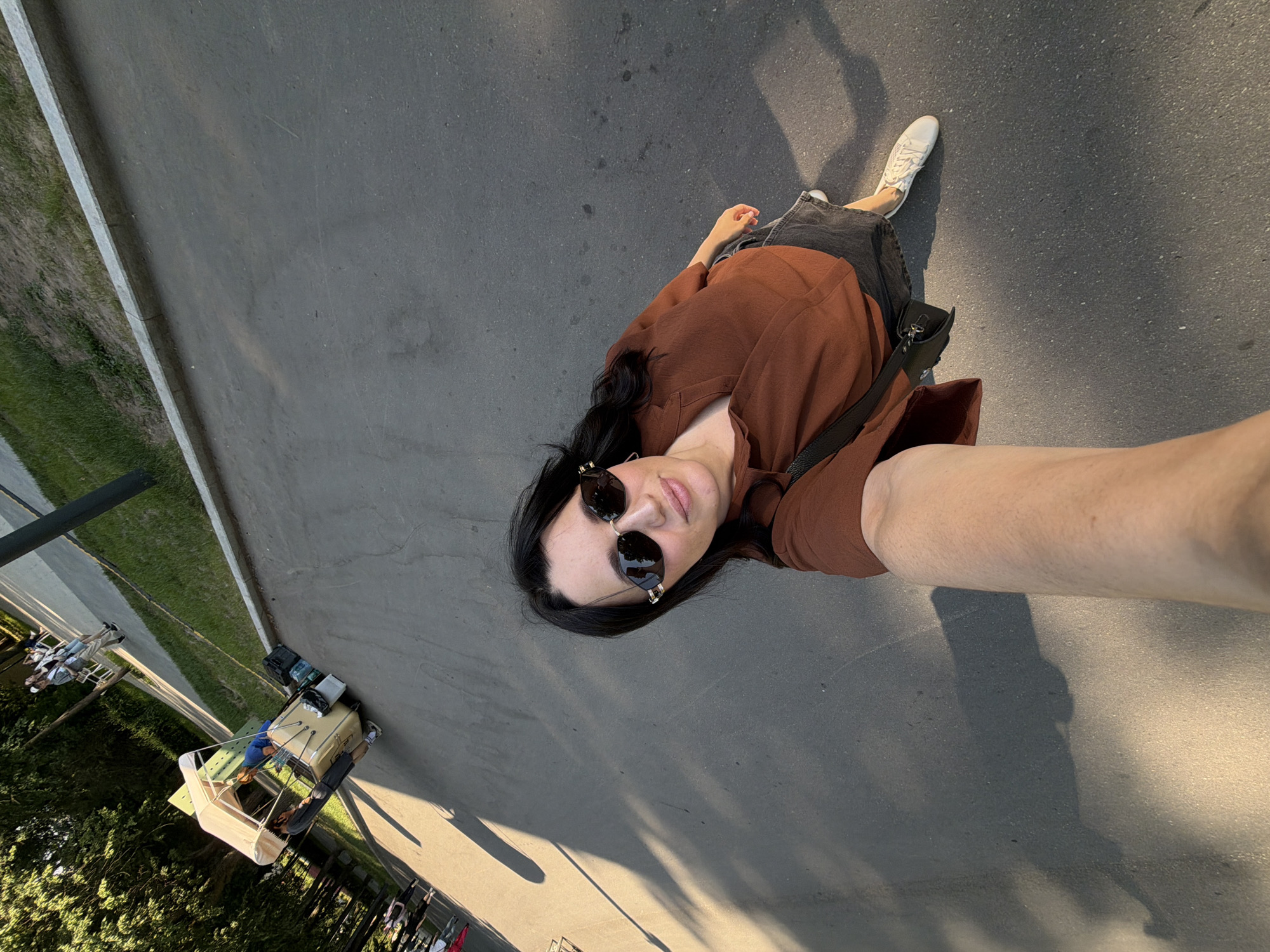 A woman is taking a selfie in an outdoor urban setting. She is wearing sunglasses, a brown shirt, and a dark skirt, with white sneakers on her feet. A black purse is slung across her body. In the background, there is a vendor cart with a canopy, and several people are visible walking along a paved path. The ground is asphalt, with a grassy area and trees bordering the path. The lighting suggests it is late afternoon or early evening, with long shadows cast across the ground. The location is identified as Moscow, Russia.