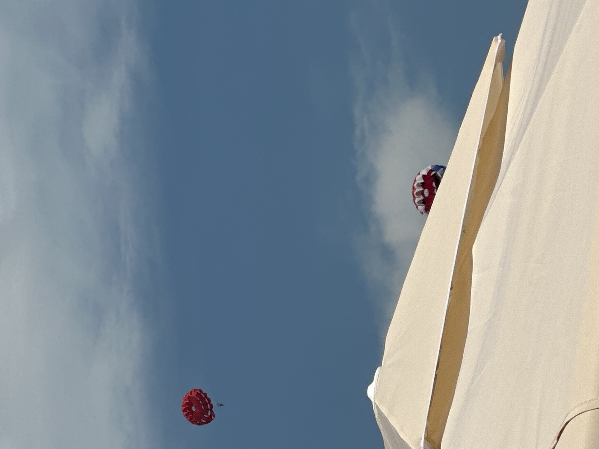 The image shows a view from beneath a large, cream-colored beach umbrella, looking up at a blue sky with some white clouds. Two parasails are visible in the sky. The first, a red parasail with white accents, is in the upper left of the frame. The second, also red and white, is closer to the horizon and partially obscured by the umbrella. The scene is likely a beach in Budva, Montenegro, given the presence of beach umbrellas and parasailing activities commonly found in tourist areas.
