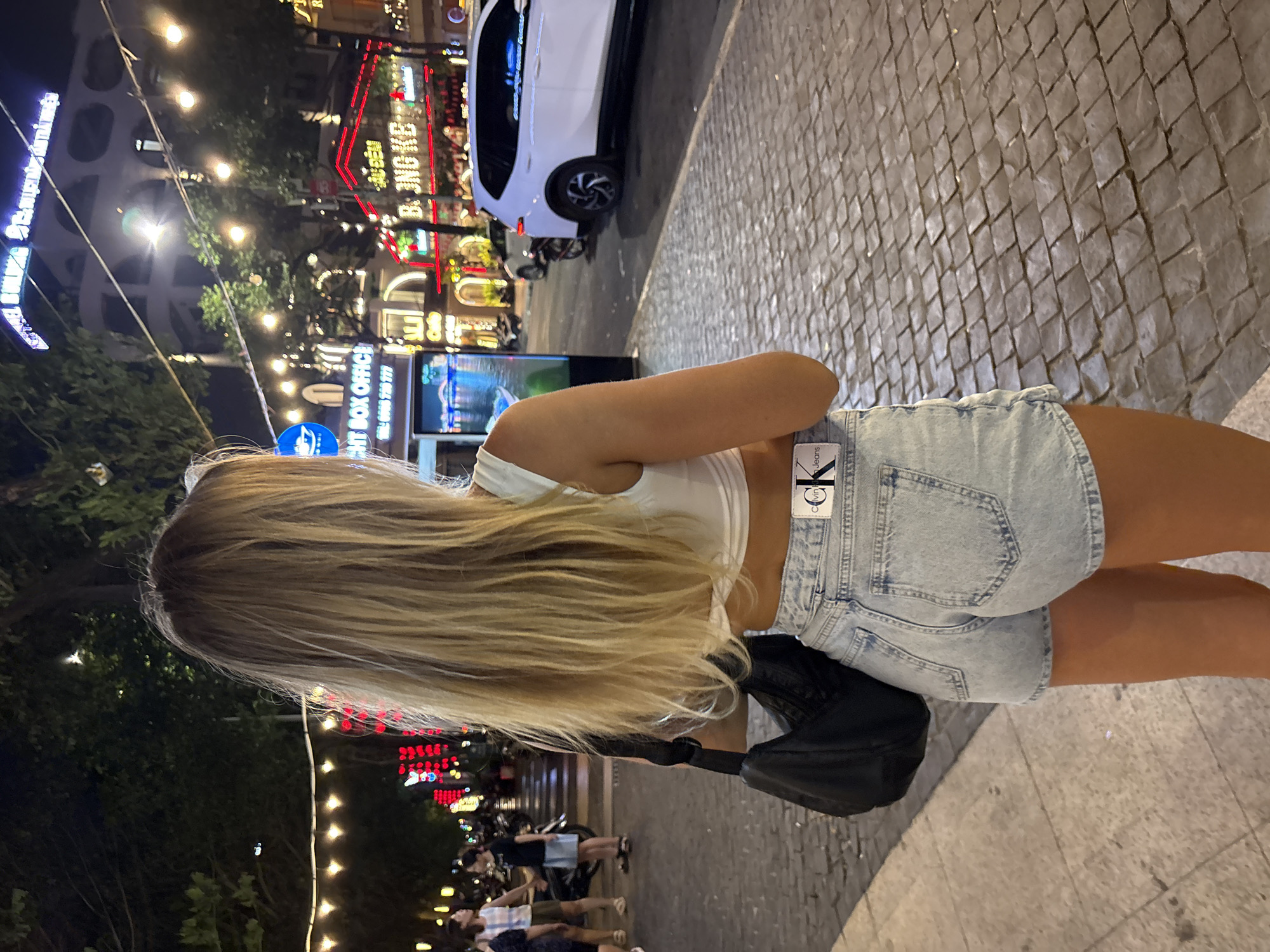 A back view of a blonde woman wearing a white tank top and denim shorts with a black purse is shown on a street in the city of Đà Nẵng, Vietnam. The backdrop consists of brightly lit buildings and streetlights. Several people are walking and there's a parked white car on the side of the road. The ground is paved with grey interlocking pavers.
