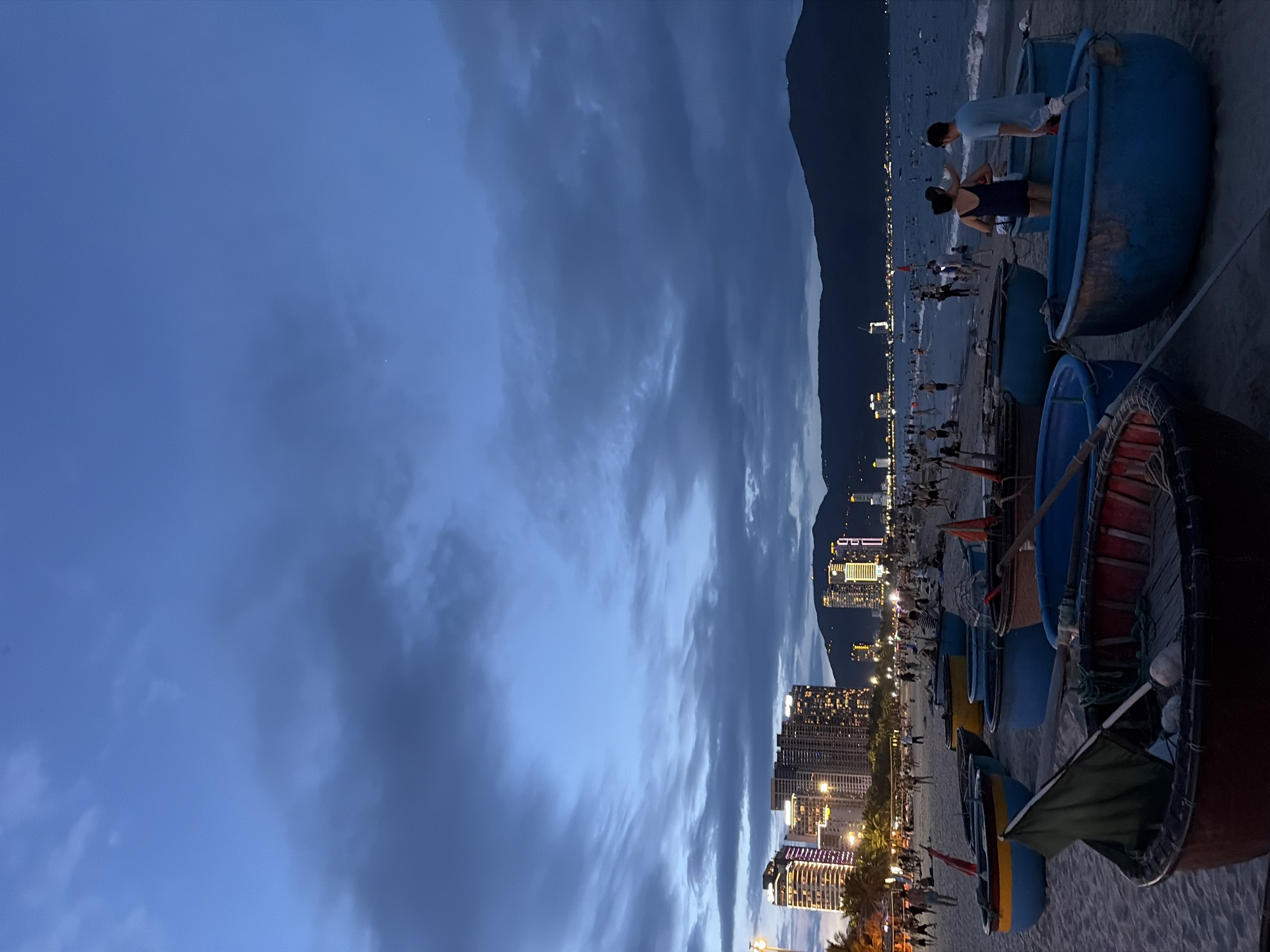 This is a vertical, medium-angle, long shot of a beach in Đà Nẵng, Vietnam, at dusk. The sky is a deep blue with heavy, dark clouds. In the distance, the city skyline is illuminated by numerous lights, with tall buildings and mountains visible. A beach is in the foreground with many round, basket-like boats on the sand. Some people are on the beach, walking and interacting, and several are in the water. The ocean has gentle waves.