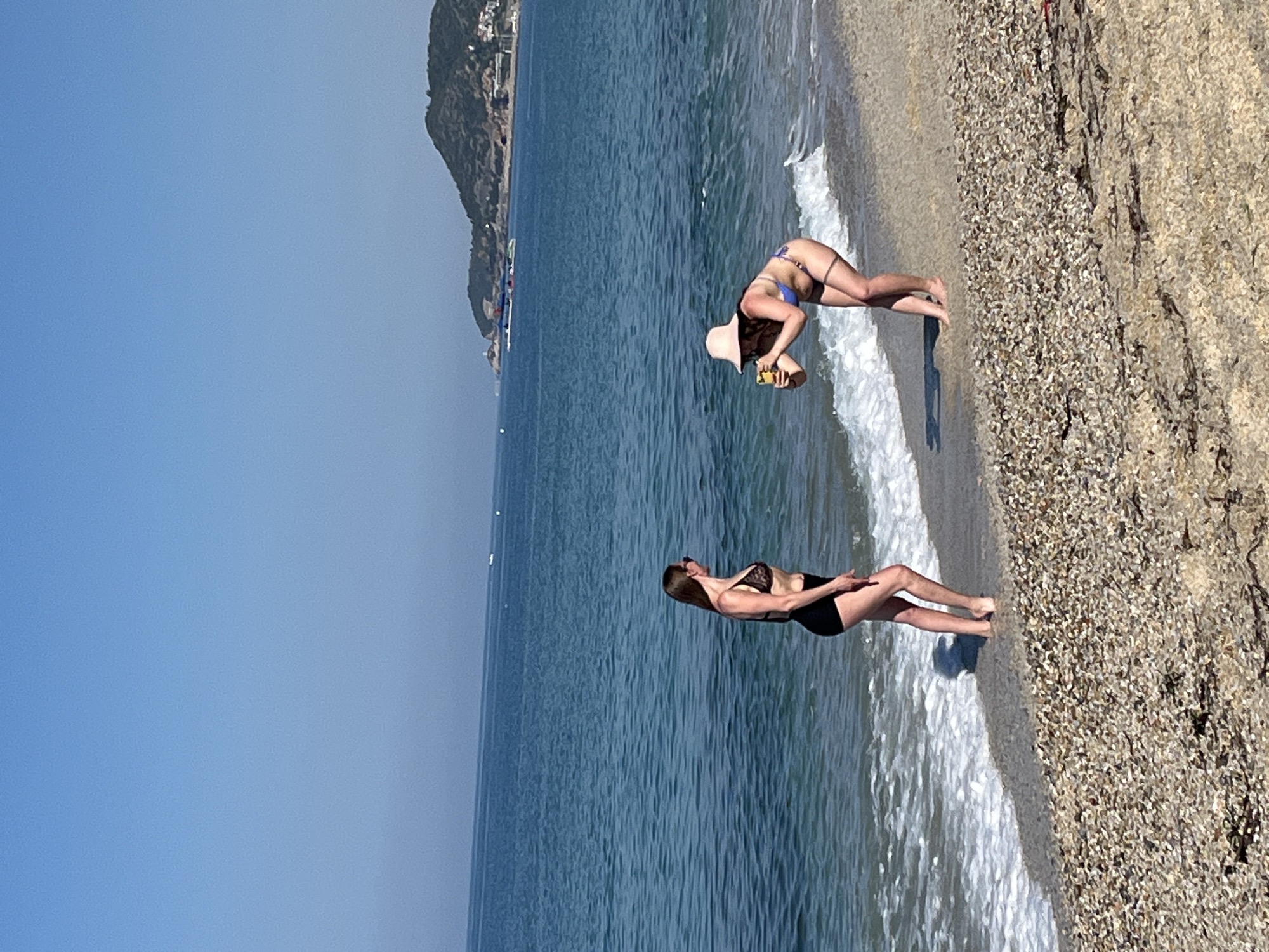 Two women are on a beach in Alanya, Turkey. The beach has small stones and is next to a blue sea under a clear blue sky. One woman, wearing a black bathing suit, is standing and looking at the water. The other woman, wearing a blue bathing suit and a hat, is bent over, possibly taking a picture or picking something up. There is a mountain in the background.