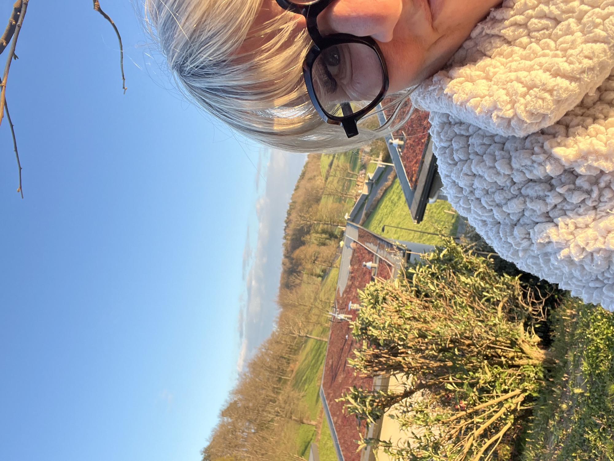The image, possibly a selfie, depicts a woman with blonde/gray hair and glasses, partially in frame. The background reveals an outdoor scene in Chambœuf, France. It includes a clear blue sky, trees (some bare), a green grassy area, and rooftops, suggesting a residential or suburban setting. The presence of the woman suggests she is outdoors, possibly on a balcony or elevated position, given the perspective.