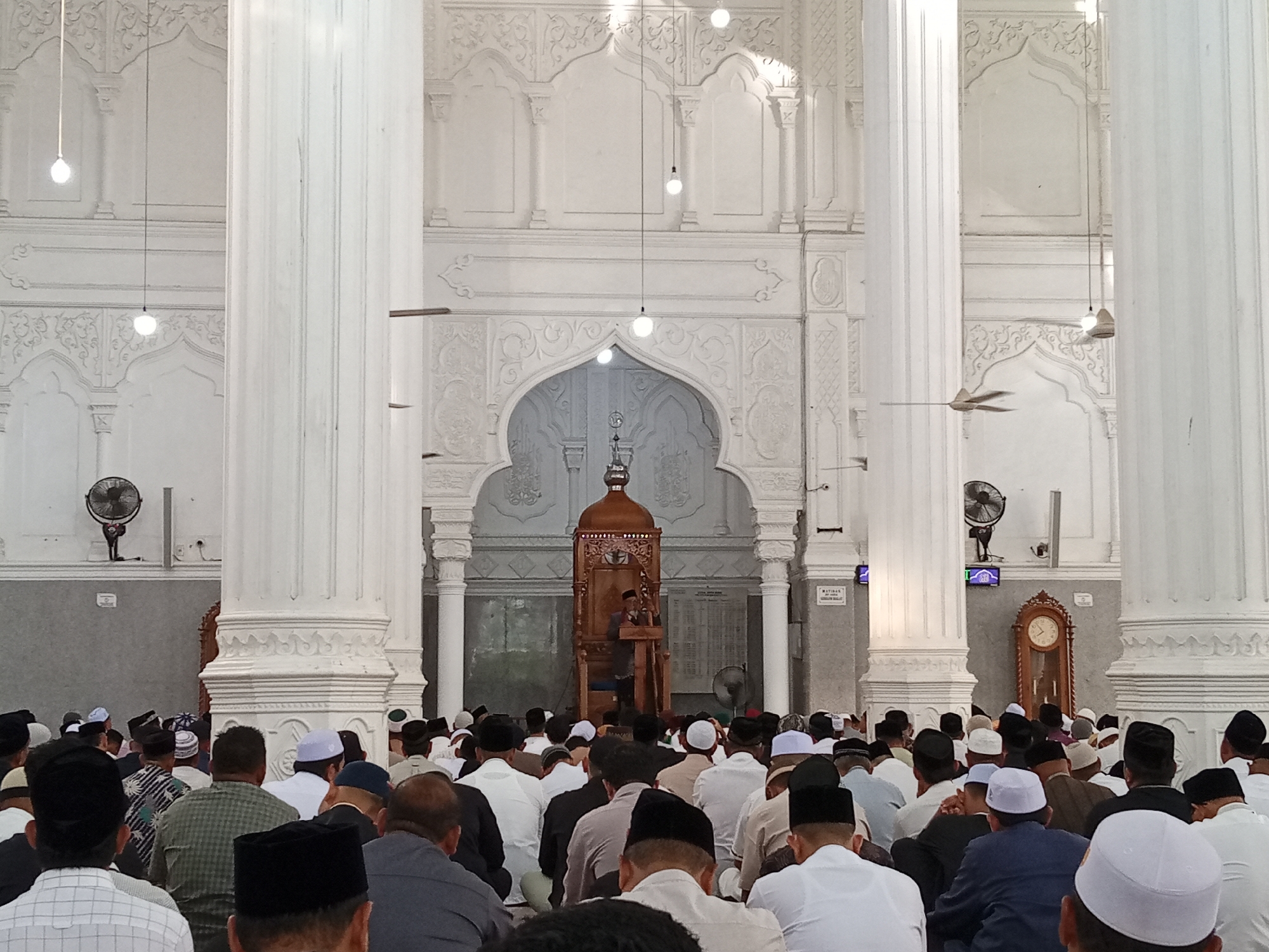 The image shows a gathering of people inside what appears to be a mosque, specifically the Islamic Center Lhokseumawe Mosque in Lhokseumawe, Indonesia. The interior features white pillars, ornate archways, and hanging light fixtures. A wooden minbar (pulpit) is visible towards the front, where a person seems to be giving a sermon or leading prayers. The congregation is composed of many men, most of whom are wearing traditional Muslim caps. They are sitting on the floor, facing the minbar, presumably engaged in prayer or listening to the speaker. Wall-mounted fans and a grandfather clock are also noticeable in the background.