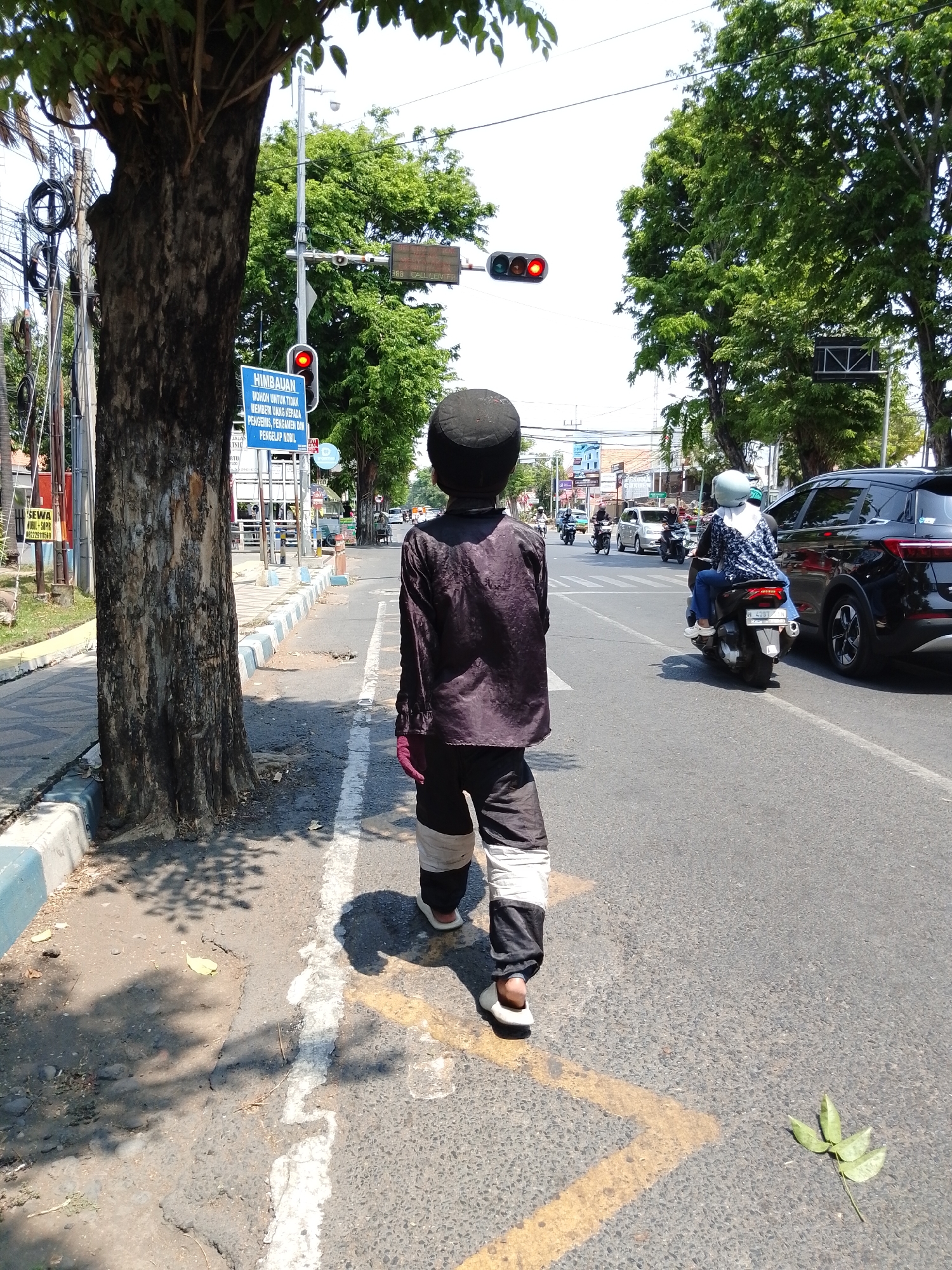 In Pasuruan, Indonesia, a person wearing a dark long-sleeved shirt and black and white striped pants walks on the right side of a paved road. The person has a dark cap on their head and is also wearing light-colored shoes. In the background, a traffic light displays a red signal. Several trees line the street, and electrical wires are visible overhead. To the right, two people are riding a scooter. A black SUV is parked or moving on the road, and other vehicles are present further down the street. The scene appears to be a sunny day in an urban or suburban area.