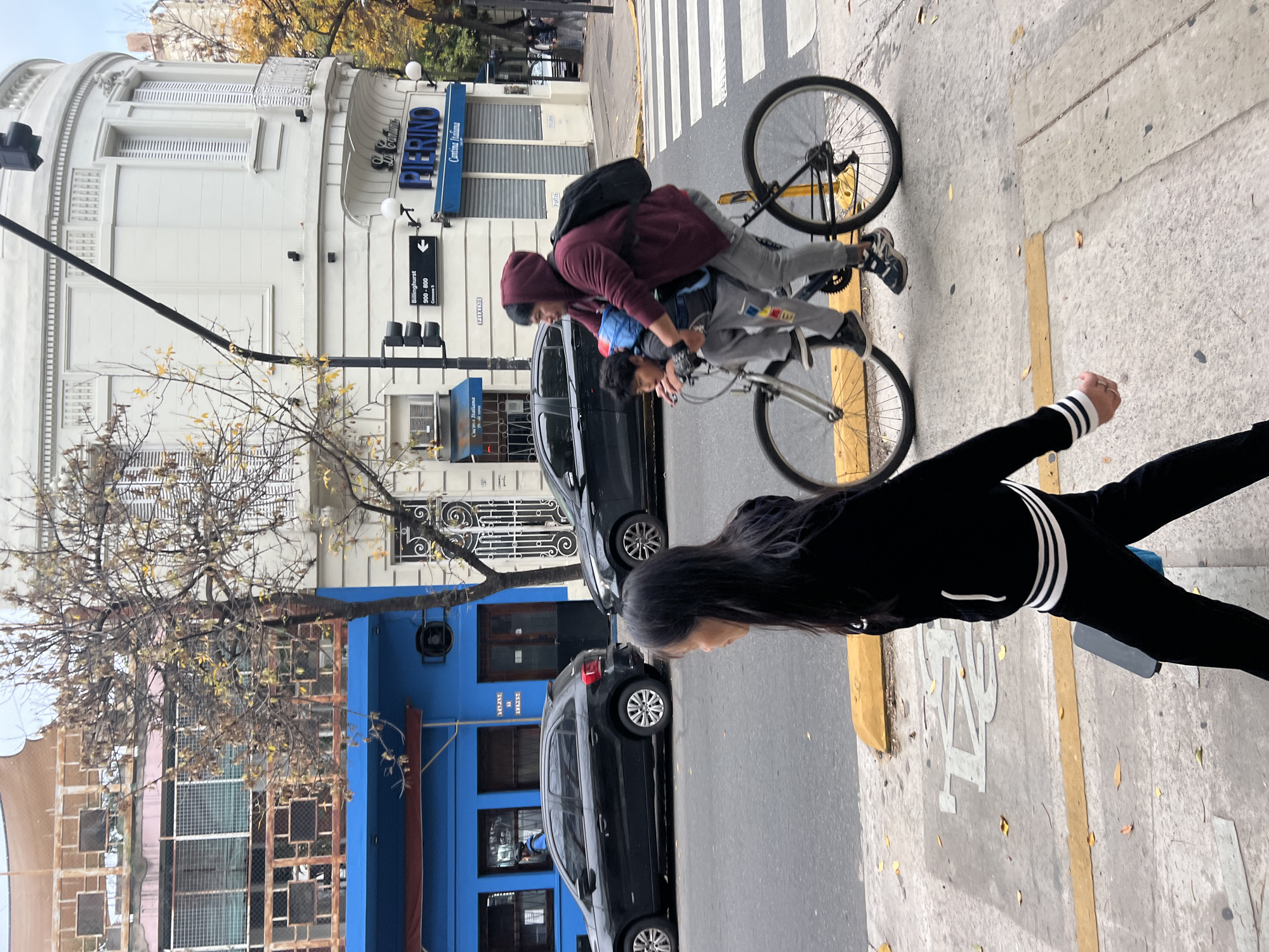 The image depicts a street scene in Buenos Aires, Argentina. A bicycle with two young males, one sitting on the handlebars, is on the street adjacent to a crosswalk. A woman is standing on the sidewalk. Parked cars are visible along the street. Buildings line the street; one is blue and another is white, with the name "Pierino" displayed on a sign. The street sign indicates Billinghurst. A bare tree is located by the blue building. The weather appears mild, possibly autumn.