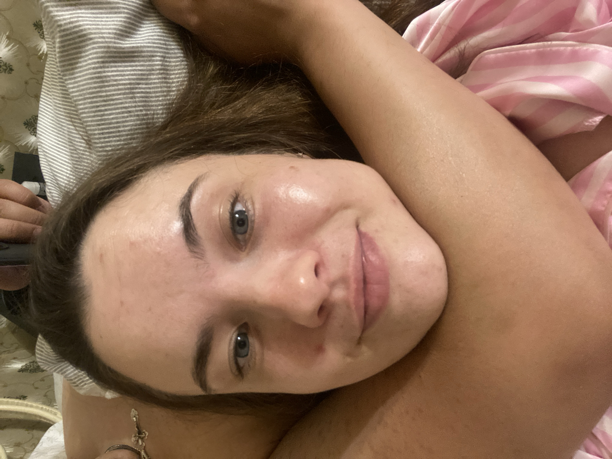 The image shows a close-up of a woman lying in bed. She has long brown hair and blue eyes, and she is looking directly at the camera with a slight smile. Her face is illuminated, highlighting her skin. An arm, presumably belonging to another person, is wrapped around her neck, and another arm is visible on the right side of the image. The woman is wearing a pink and white striped garment. The background consists of a pillow with gray and white stripes and a patterned bedspread. There are no discernible features that place this scene in Poltava, Ukraine, or any specific city.