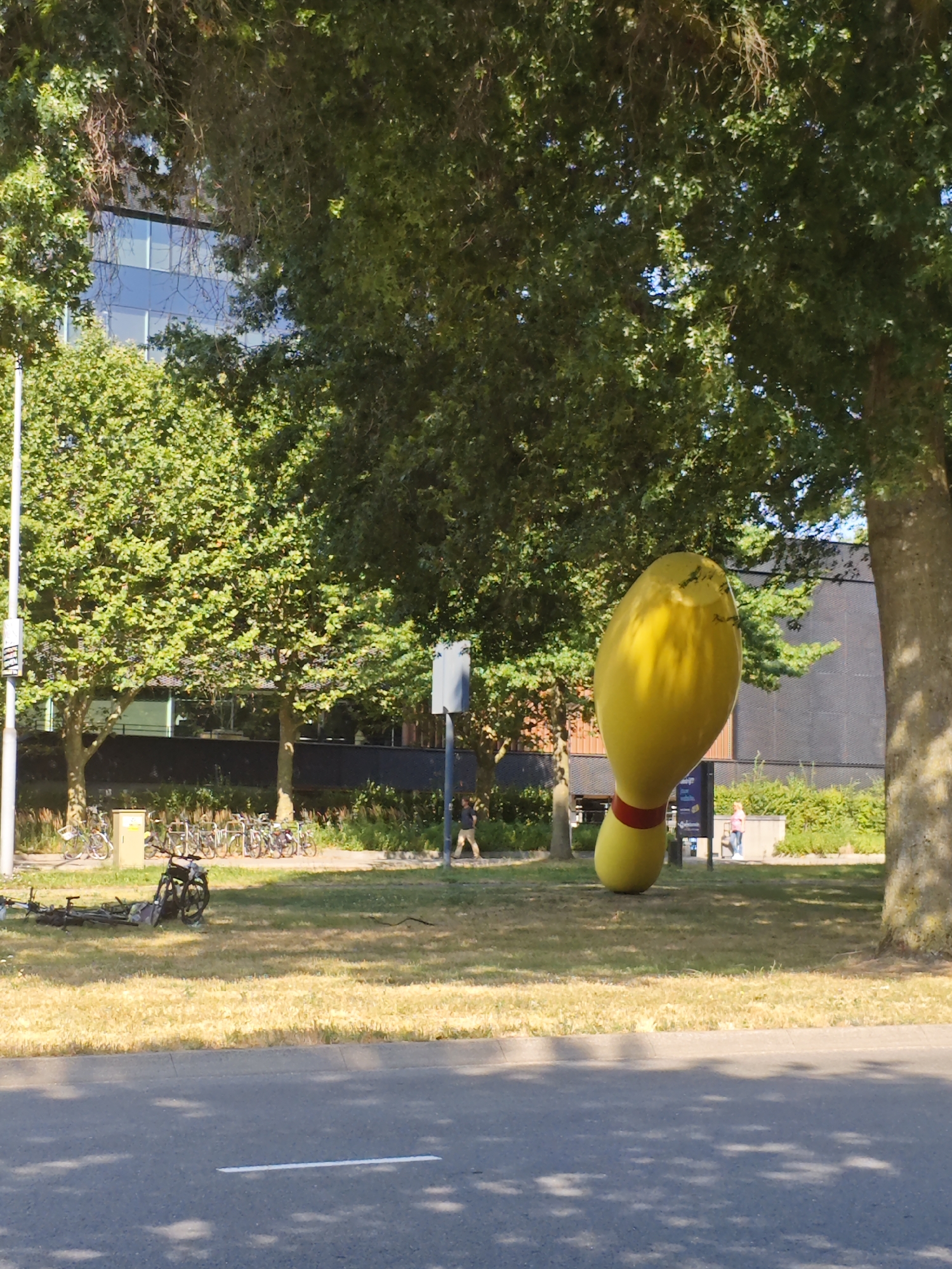 The image shows a sunny outdoor scene in Eindhoven, Netherlands, featuring a large, bright yellow sculpture shaped like a bowling pin, complete with a red band around its neck. The sculpture stands prominently on a grassy area, partially shaded by lush green trees with dappled sunlight falling on the grass. In the background, a modern building with a dark facade and many windows is visible, along with a row of parked bicycles. A fallen bicycle lies on the grass in the foreground. Several people are present in the scene, including one walking in the background near the parked bicycles, another walking past the sculpture, and a person near the building in the far background. The foreground shows a paved road with a white line, indicating it is likely a street or sidewalk. The overall impression is a vibrant, urban park setting.