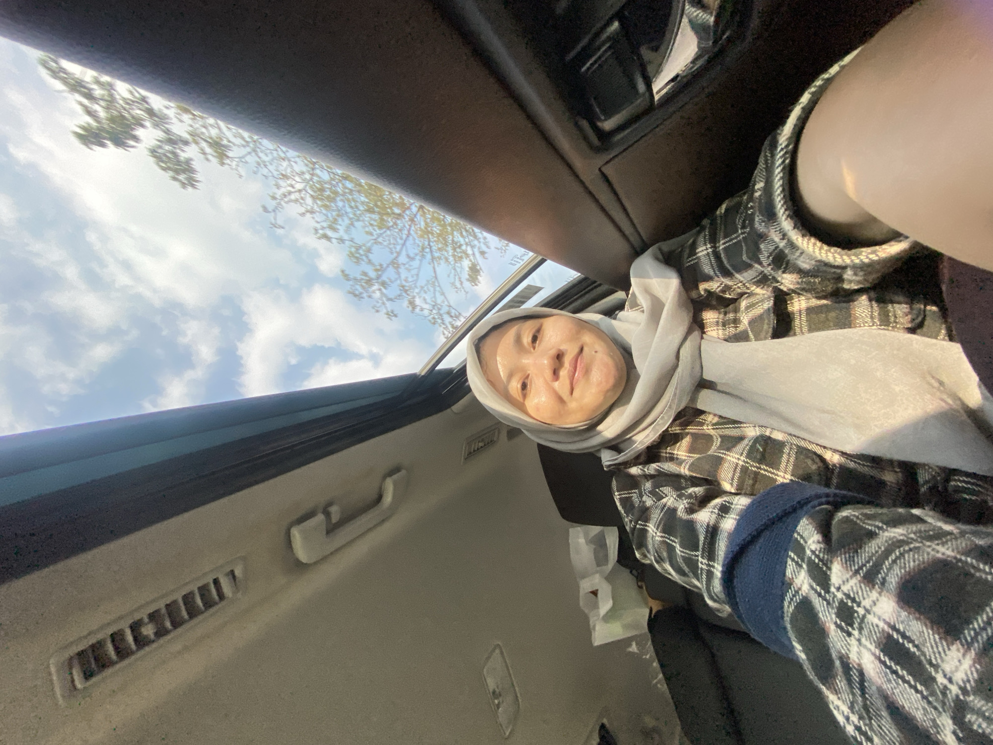 A woman wearing a light gray hijab and a black and white plaid shirt is inside a car. She is smiling and looking at the camera. The car has a sunroof open, revealing a blue sky with white clouds and some trees in the background. The interior of the car, including the ceiling with air vents and a handle, is visible. The scene appears to be in Mandar Sari, Indonesia, given the natural lighting and foliage.